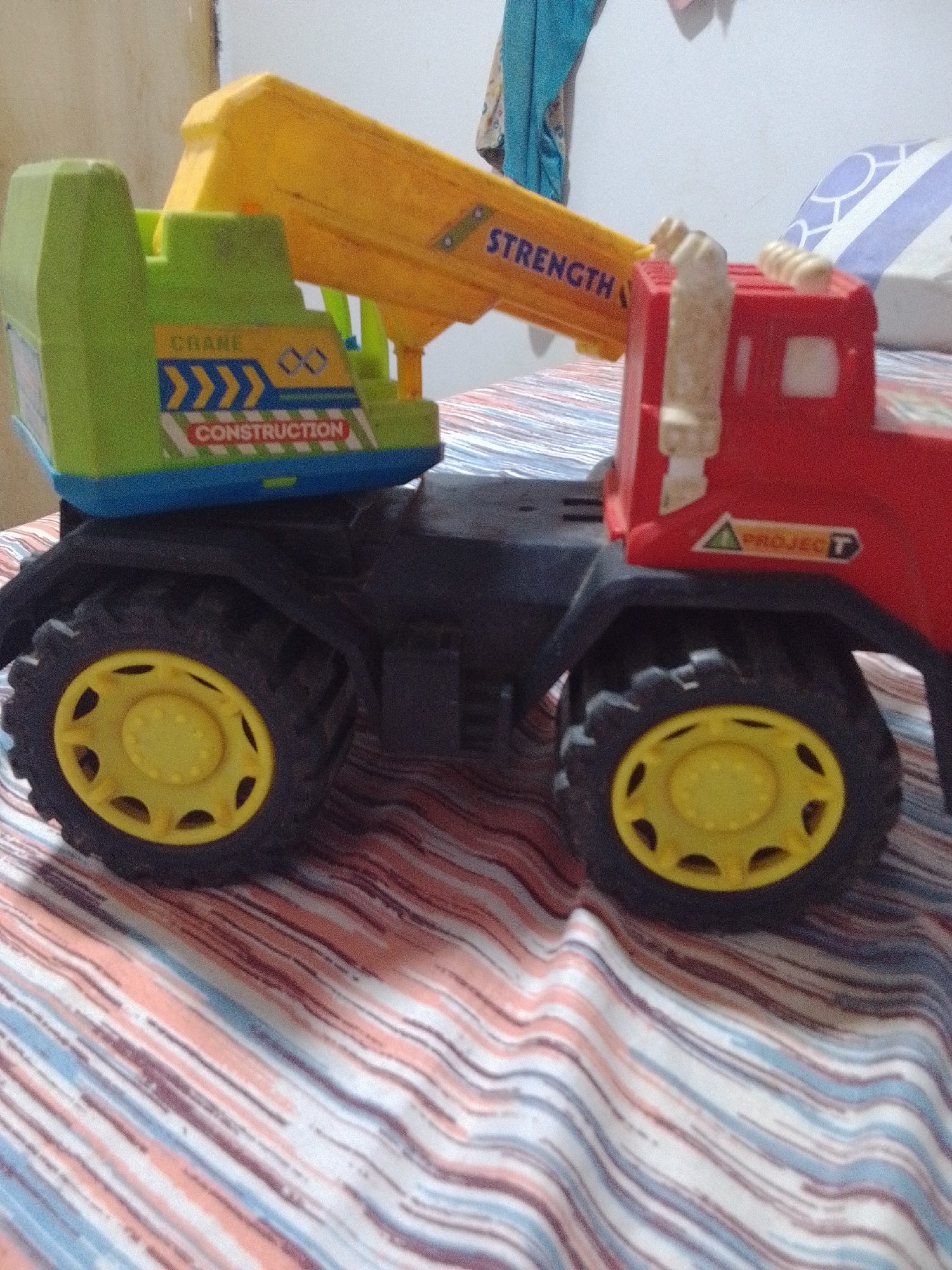 A toy crane truck is displayed on a bed with striped sheets. The truck is red and green with yellow accents, featuring a crane arm that extends from the top. The crane arm has the word "STRENGTH" printed on it in blue letters. The truck's body is labeled "CRANE CONSTRUCTION" in blue and red lettering. The truck has large black wheels with yellow rims. A sticker with a green triangle and the word "PROJECT" in red is visible on the side of the red cab. The background is blurred, showing a white wall and a portion of a patterned pillow. The setting appears to be indoors, possibly a bedroom, given the presence of the bed and wall. No people are visible in the image.