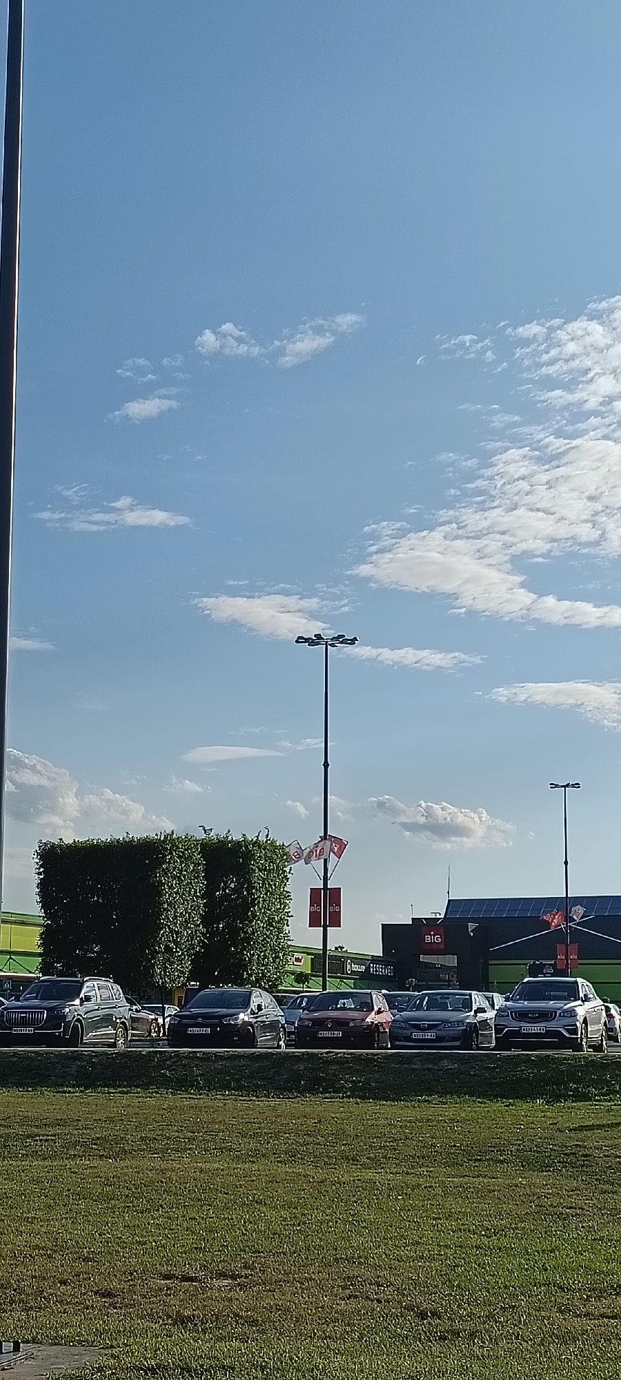 This is a vertical, eye-level shot taken outdoors on a clear, sunny day, likely in the late afternoon given the low angle of the sun casting soft shadows. The scene depicts a parking lot in front of a commercial building, possibly a shopping center, in Novi Sad, Serbia.

In the foreground, a well-maintained, green grassy area slopes gently down towards the parking lot. The parking lot itself is filled with several cars parked in rows. Among the visible vehicles are a dark grey SUV on the far left, a black sedan, a red hatchback, a grey sedan, and a white SUV on the far right, with other cars partially visible behind them. The cars appear to be newer models.

Behind the parking lot, there are some notable architectural and landscape features. Two large, rectangular, meticulously trimmed green hedges stand prominently in the mid-ground. To the right of the hedges, a tall lamppost with multiple light fixtures rises into the bright blue sky, which is dotted with scattered white clouds. Adjacent to the lamppost and behind the hedges, a building with a dark facade is visible. This building features a large "BIG" logo, a common branding for shopping centers. Further to the right, another part of the commercial building has a green facade and a sloped, dark-patterned roof, possibly indicating solar panels. Several flags, including those with "BIG" branding and possibly country flags, are displayed on poles near the building.

The overall impression is one of a modern commercial area on a pleasant day. There are no people visible in the frame, suggesting a time when the area is less busy or the photo was taken from a distance. The scene conveys a sense of everyday activity in a retail or entertainment district.