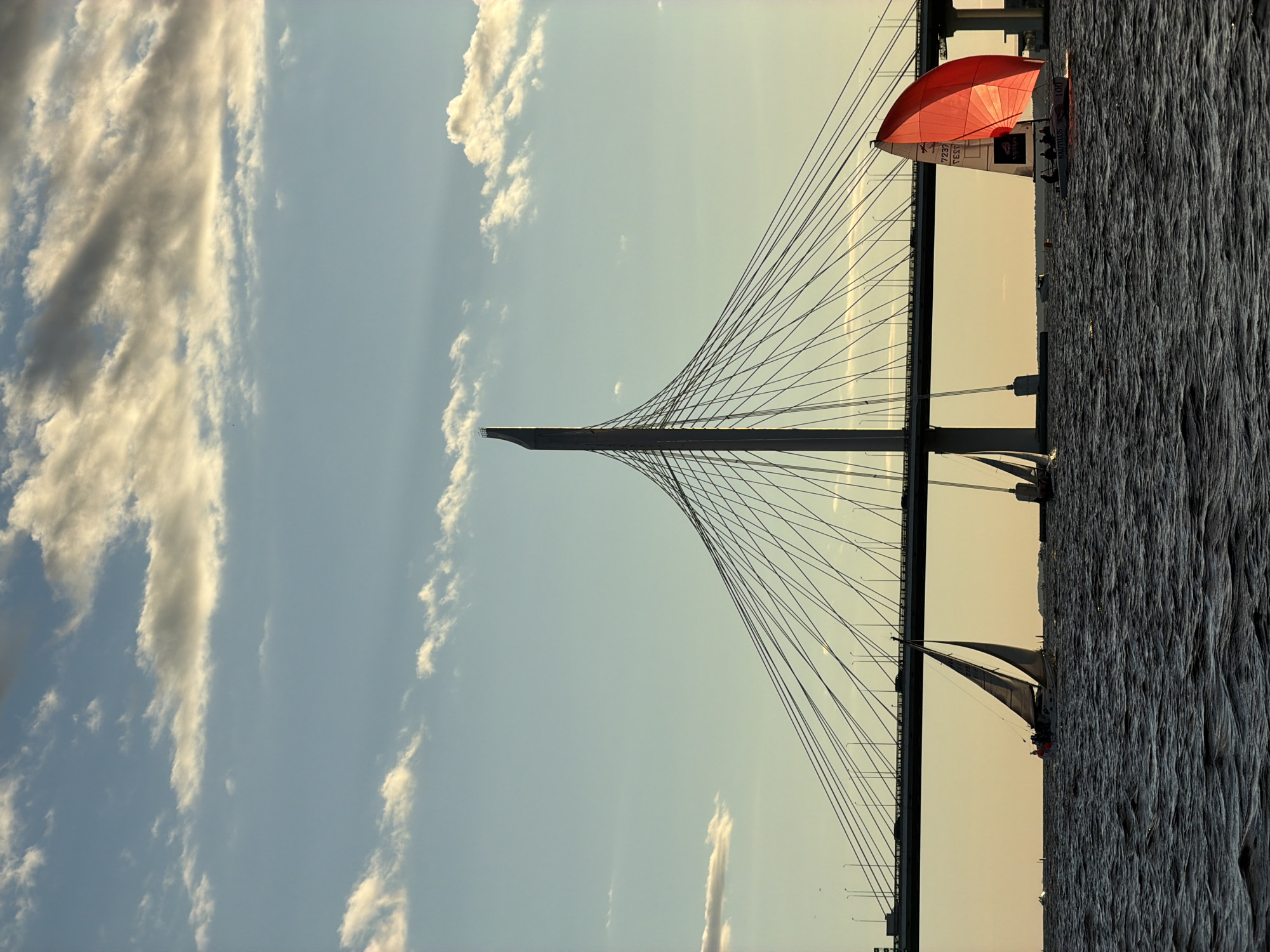 The image depicts a seascape in Saint Petersburg, Russia, featuring sailboats and a large cable-stayed bridge. The sky is a pale blue with scattered white and grey clouds, illuminated by the warm glow of sunset. The water is dark and choppy, reflecting the subdued light.
In the foreground, several sailboats are visible. One prominent sailboat on the right has a bright red sail, emblazoned with the word "NAUTILUS" and the number "100." Several figures are discernible on this boat. To the left, a larger sailboat with two white sails is seen, also with people on board. Other sailboats are present further in the background.
The central focus of the image is the massive cable-stayed bridge, spanning across the water. Its towering central pylon, a distinctive architectural feature, rises high into the sky, supporting a web of cables that stretch across the roadway. Cars can be seen driving on the bridge, silhouetted against the lighter sky. The overall atmosphere is one of serene maritime activity set against a backdrop of impressive urban infrastructure.