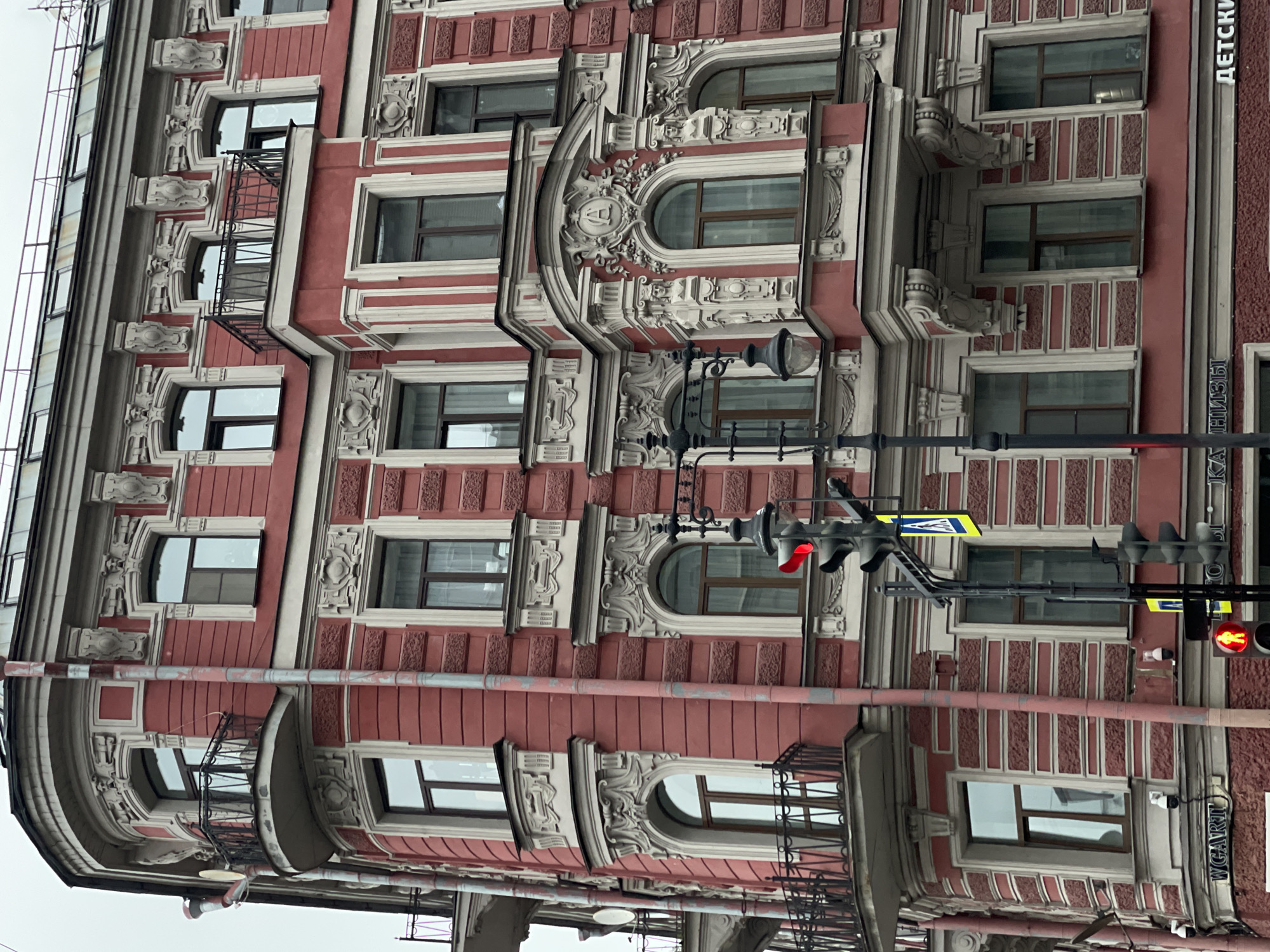 The image captures a corner of a building in Saint Petersburg, Russia, characterized by its distinct architectural style. The facade is primarily red with white trim and ornate details around the windows, some of which are arched. The building features multiple windows on each visible floor, and some windows have small balconies with black iron railings. A traffic light with red lights illuminated is attached to a pole in front of the building, along with a pedestrian crossing sign. The sky is overcast, contributing to a muted overall color palette. Russian text is visible on the building's side, indicating the location is indeed in Russia.