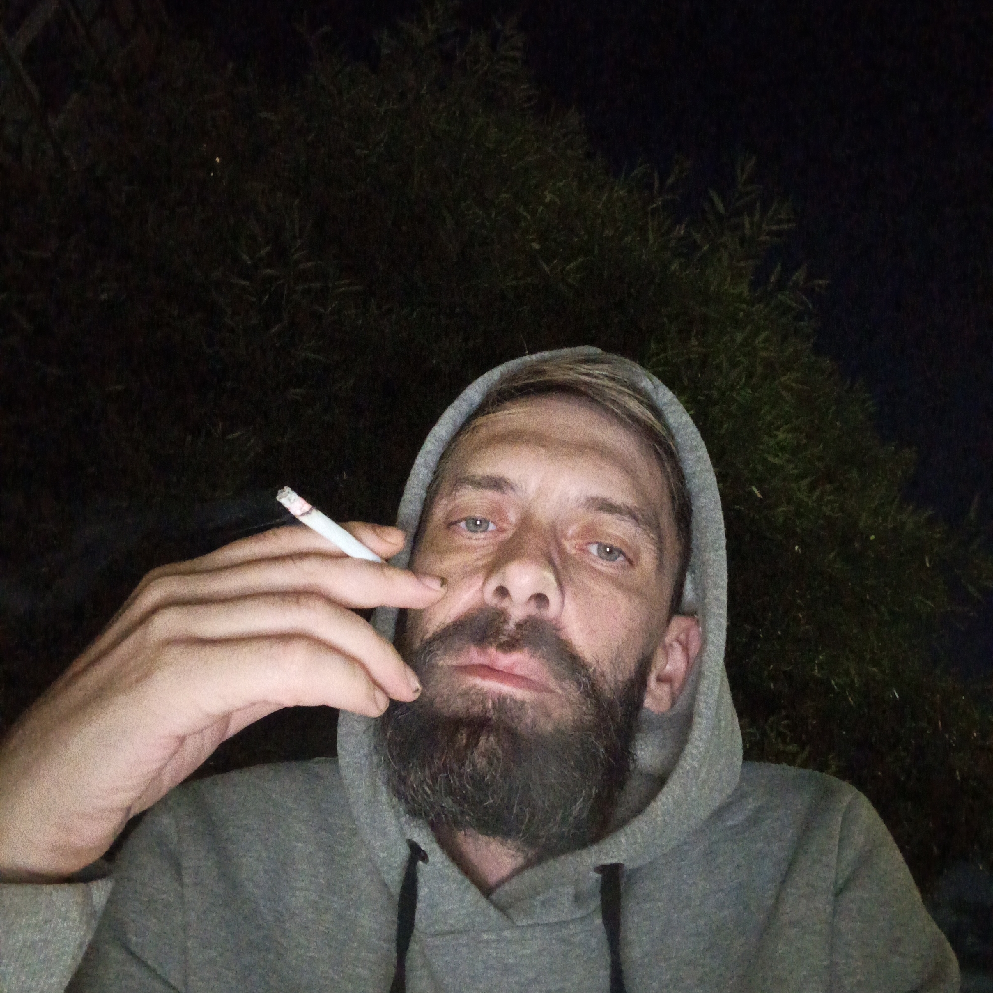 A close-up, eye-level, waist-up, selfie-style shot shows a man with a beard and blonde hair wearing a grey hooded sweatshirt. He is holding a lit cigarette in his right hand, bringing it to his mouth. His gaze is directed towards the camera. The background consists of dark foliage, suggesting an outdoor nighttime setting. The image is set in Markovskiy, Russia.