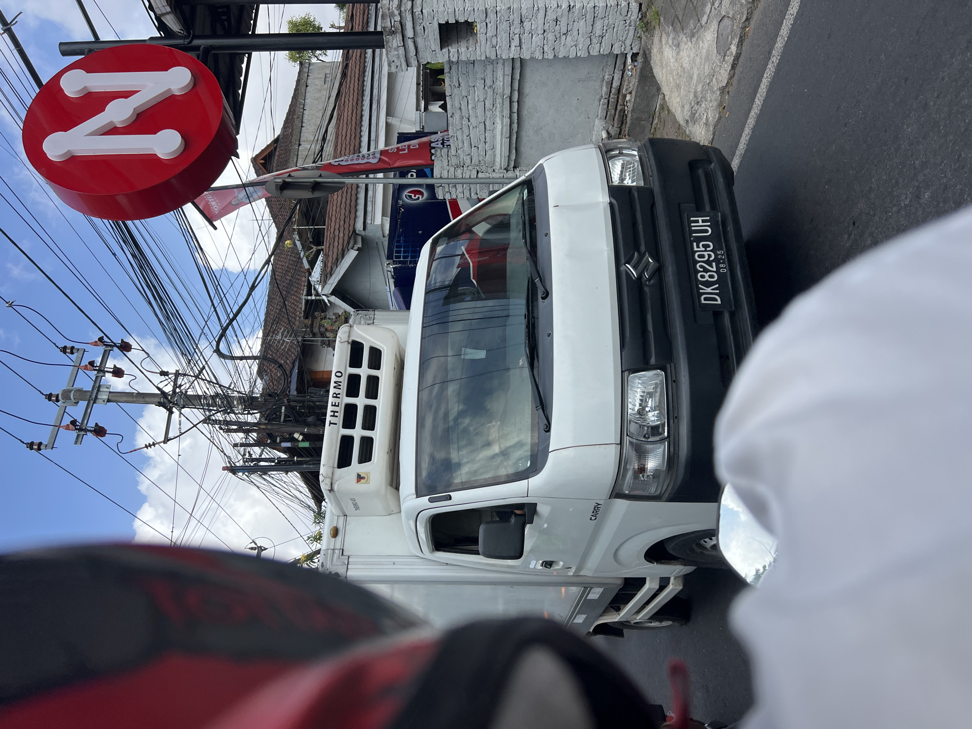 The image shows a white Suzuki Carry truck driving on a street in Denpasar, Indonesia. The truck has a refrigeration unit on top and displays the license plate DK 8295 UH. In the background, there is a building with a red circular sign that has a white "N" logo. There are also power lines and some residential buildings visible. The perspective is from someone riding on a motorcycle behind the truck, with a portion of their white shirt and red helmet visible in the foreground. The sky is blue with some clouds.