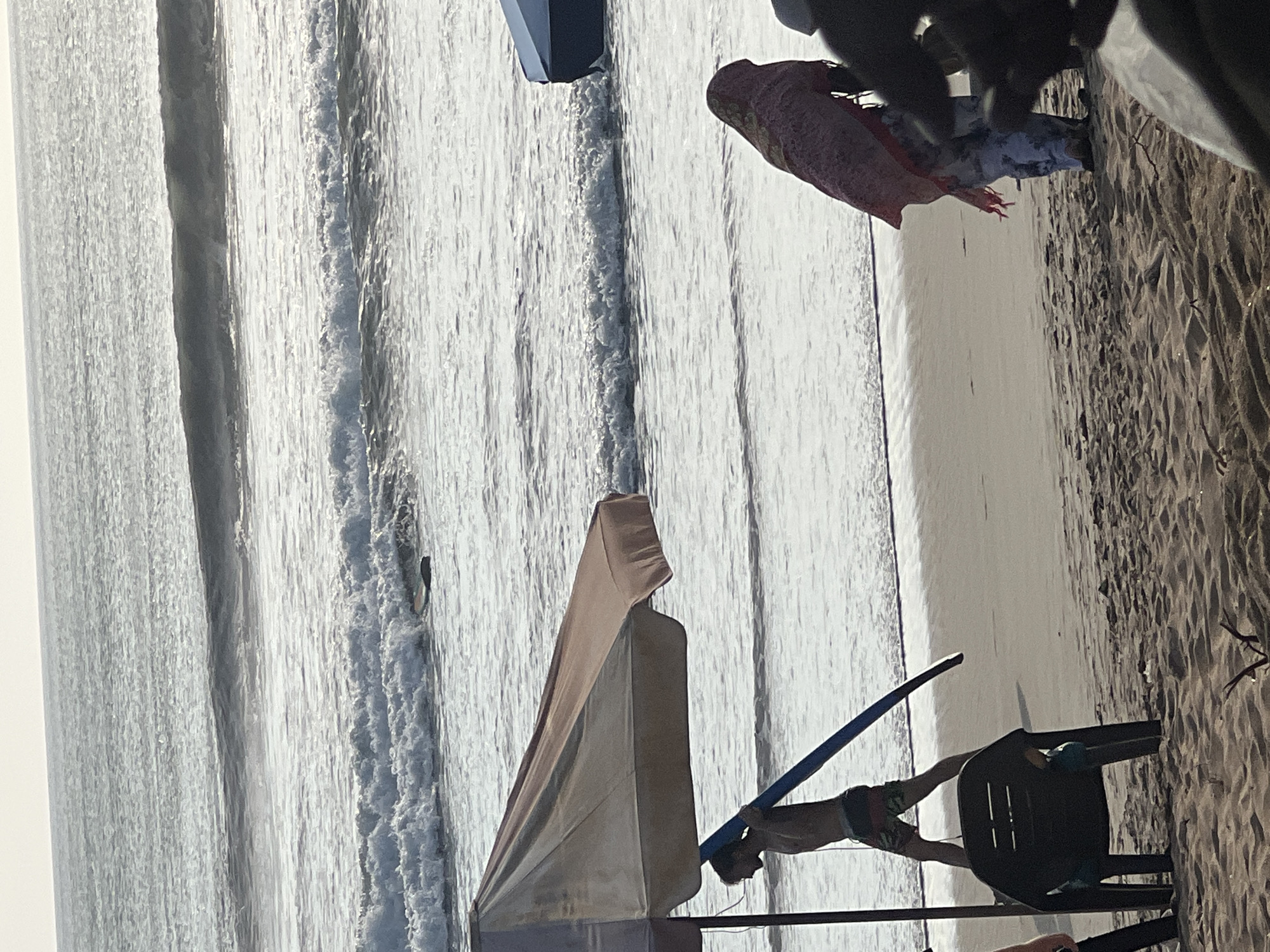 The image depicts a beach scene in Kuta, Indonesia, likely during daylight hours given the illumination. In the foreground, a large, light-colored beach umbrella provides shade over a black plastic chair. To the left of the umbrella, a young person, possibly male, is shown from the waist down, wearing patterned shorts and carrying a blue surfboard horizontally under their arm, walking on the sand towards the water. Further to the right, a person is visible from behind, wearing a patterned pink headscarf and patterned trousers, standing on the beach. In the background, the ocean stretches out with moderate waves breaking near the shore. The sand on the beach appears light brown and textured. The sky is overcast and bright, suggesting diffused sunlight.
