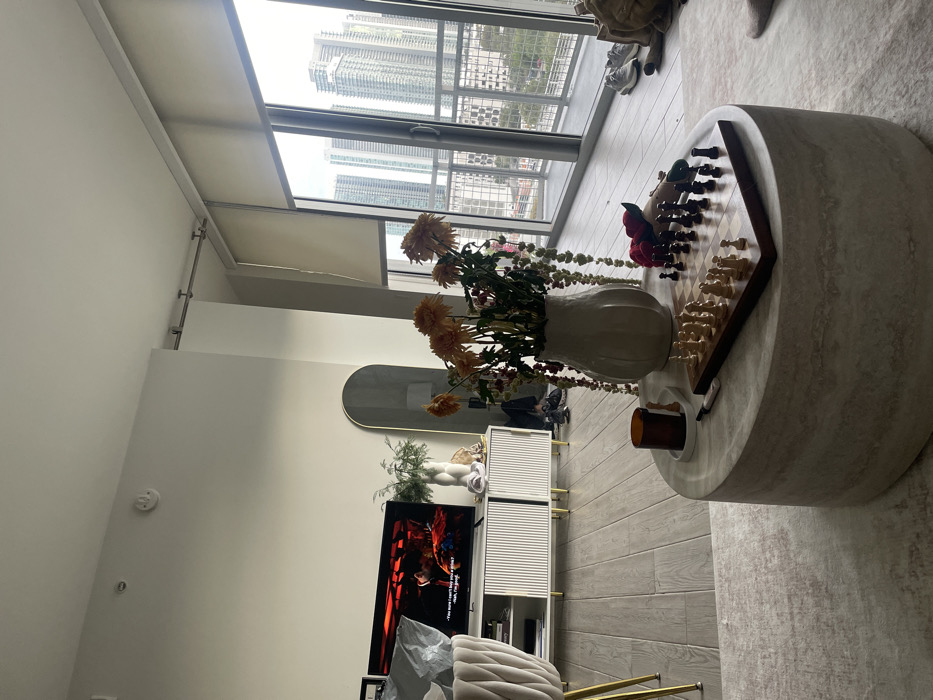 A modern apartment living room features a large window with a city view. A round marble coffee table holds a vase of dried flowers and a chessboard. Below, a television sits on a white cabinet with gold legs. A long mirror hangs on the wall. The room is decorated in neutral tones with wooden flooring and light walls.