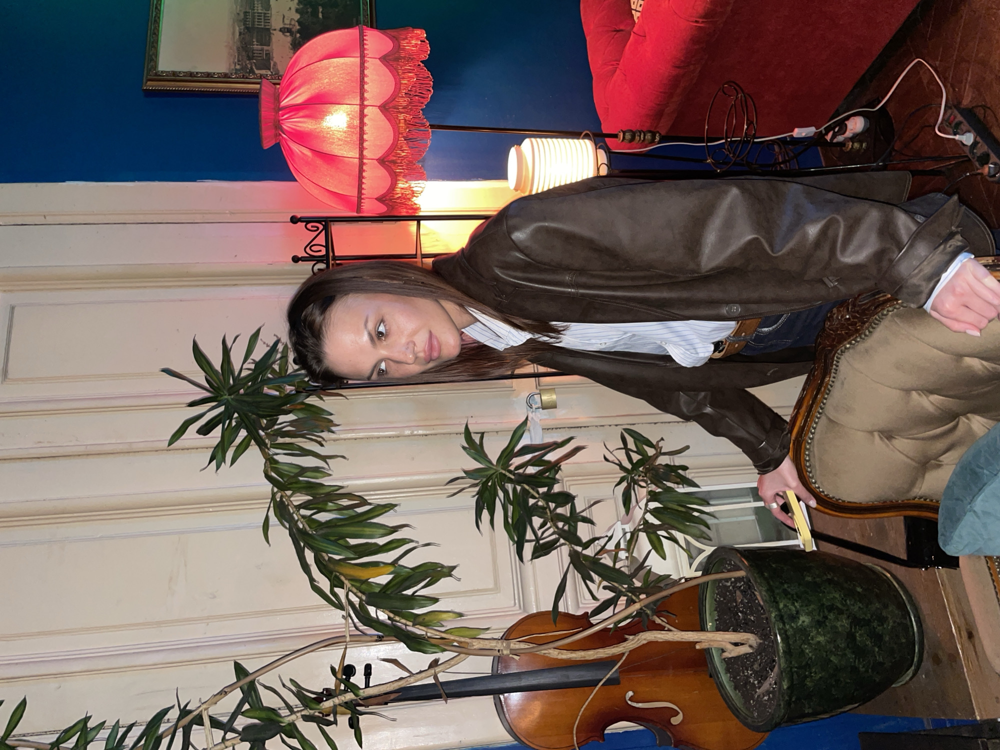 A woman in a brown leather jacket leans on an upholstered chair indoors. She is fair-skinned, with brown hair and is wearing a striped shirt underneath the jacket. The jacket sleeves are slightly rolled up, and a belt is visible at her waist. She rests her arm on the chair, holding a yellow object.
A large houseplant in a green pot is in the foreground, with a brown cello placed in front of it. The door behind the plant is white and has a lock on it. The room features a red sofa, an ornate red lamp with a pleated shade, and a smaller white lamp. A framed picture hangs on the blue wall above the lamps.
The image appears to have been taken indoors, with a mix of artificial and natural lighting. The scene includes furniture, decor, and a musical instrument, suggesting a lived-in or staged environment. This is probably in Saint Petersburg, Russia.