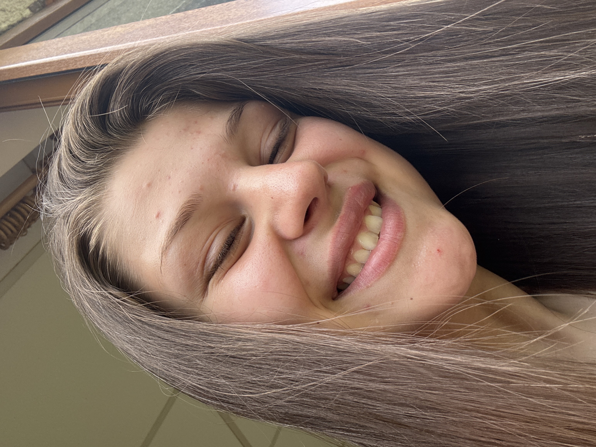 A medium close-up shot focuses on the face of a young person with their eyes closed and a wide smile. Their hair, a shade of brown with lighter highlights, is parted on the side and cascades down around their face. The person has light-colored skin with some minor blemishes visible on their forehead and cheek. The background is out of focus, appearing to be an outdoor setting with a hint of a wooden frame on the right side, suggesting a window or door. The lighting suggests it is daytime.