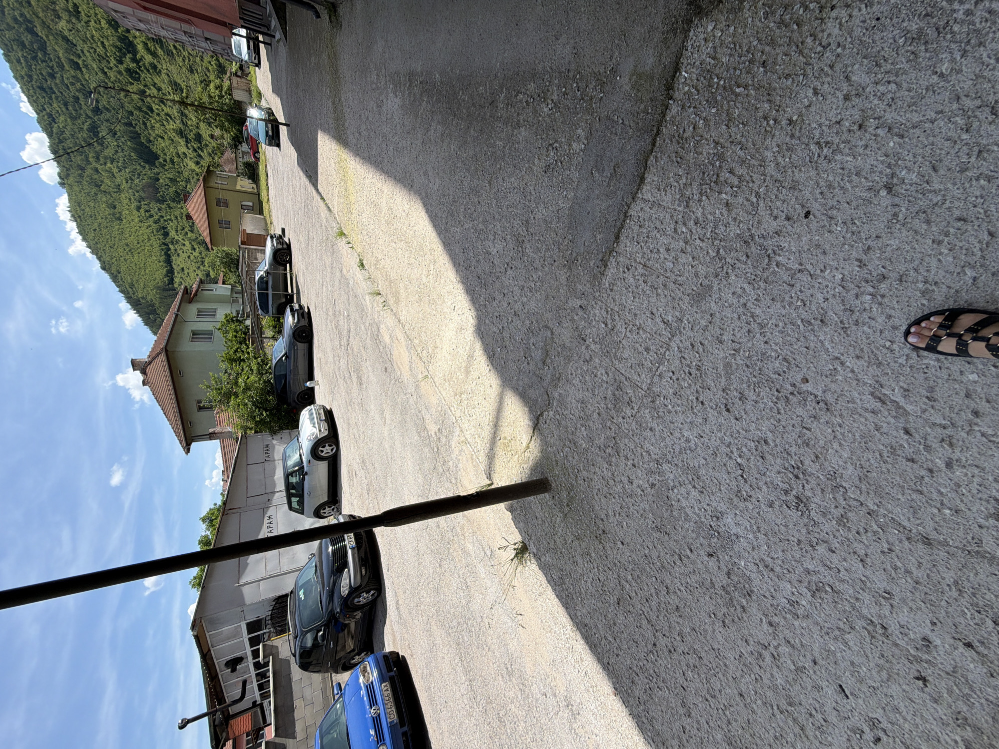 In Etropole, Bulgaria, a scene unfolds on a sunny day in what appears to be a residential area or a small commercial lot. A black car, possibly a Chrysler PT Cruiser, is parked next to a blue Volkswagen Golf. Further down, a silver Mini Cooper and a gray BMW SUV are visible. Across the lot, a dilapidated building with a slanted roof stands, bearing Cyrillic lettering that translates to "GARAGE." Residential buildings, some with red-tiled roofs, line the background, backed by a lush, tree-covered mountain slope. A person's sandal-clad foot is in the foreground, hinting at the presence of an individual documenting the scene. The ground is a mix of cracked asphalt and gravel, casting long shadows from a nearby pole.