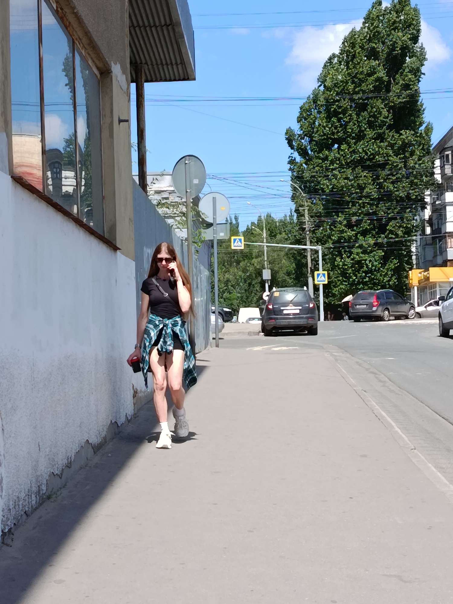 A woman with long brown hair, wearing sunglasses, a black t-shirt, shorts, white socks and sneakers, and a green and blue plaid shirt tied around her waist, walks on a sidewalk next to a street. She is holding a red cup in her left hand and talking on her phone. A white wall is to her left, and a street with several cars is to her right. The sky is blue with some white clouds. The scene appears to be in the city of Saratov, Russia, based on the architecture and vegetation.