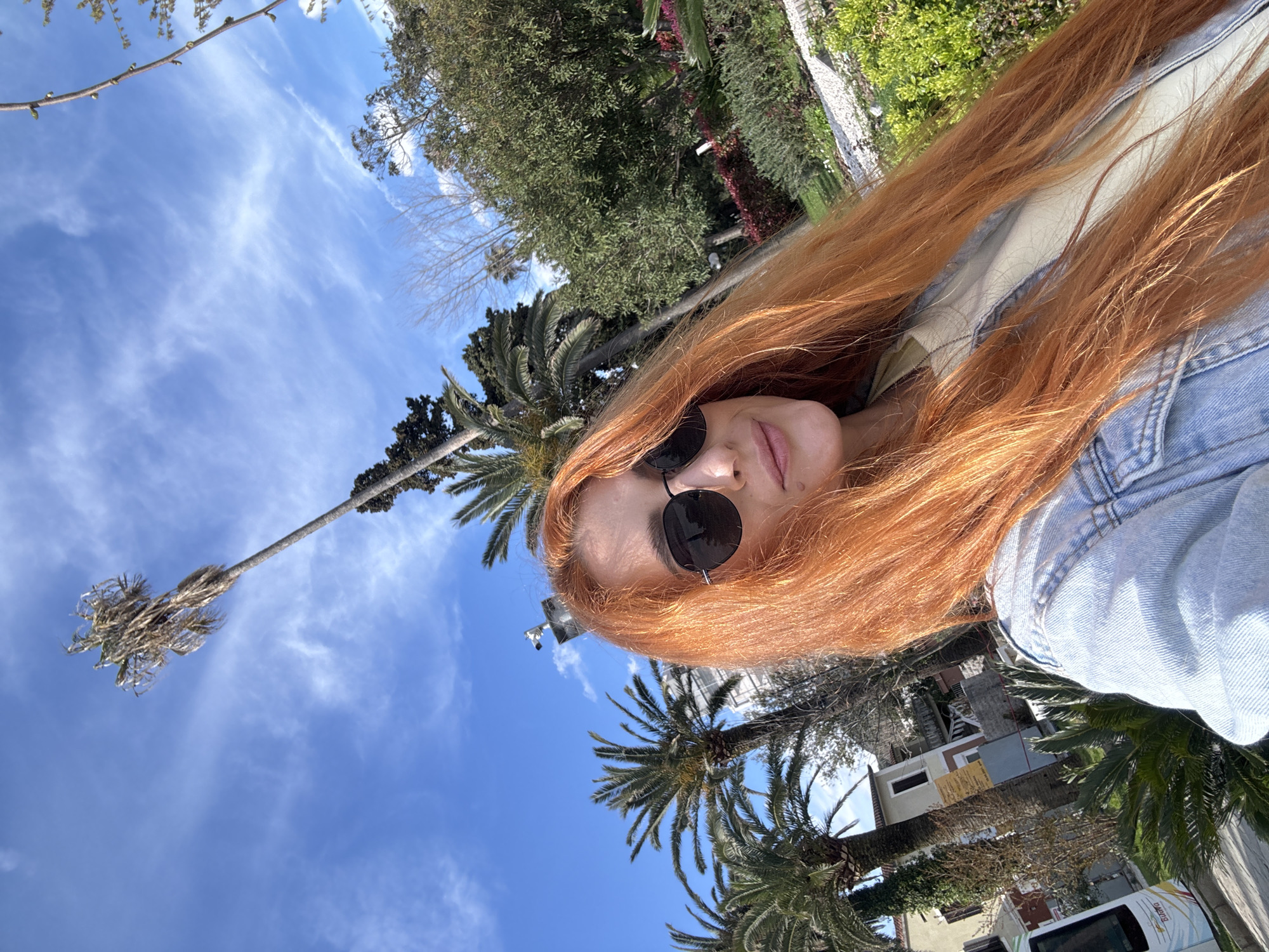 A selfie taken in Budva, Montenegro, shows a woman with long red hair and sunglasses. She is wearing a denim jacket. The background includes palm trees, a building, a white van, and a blue sky with scattered clouds. The location is likely an urban area with Mediterranean vegetation.