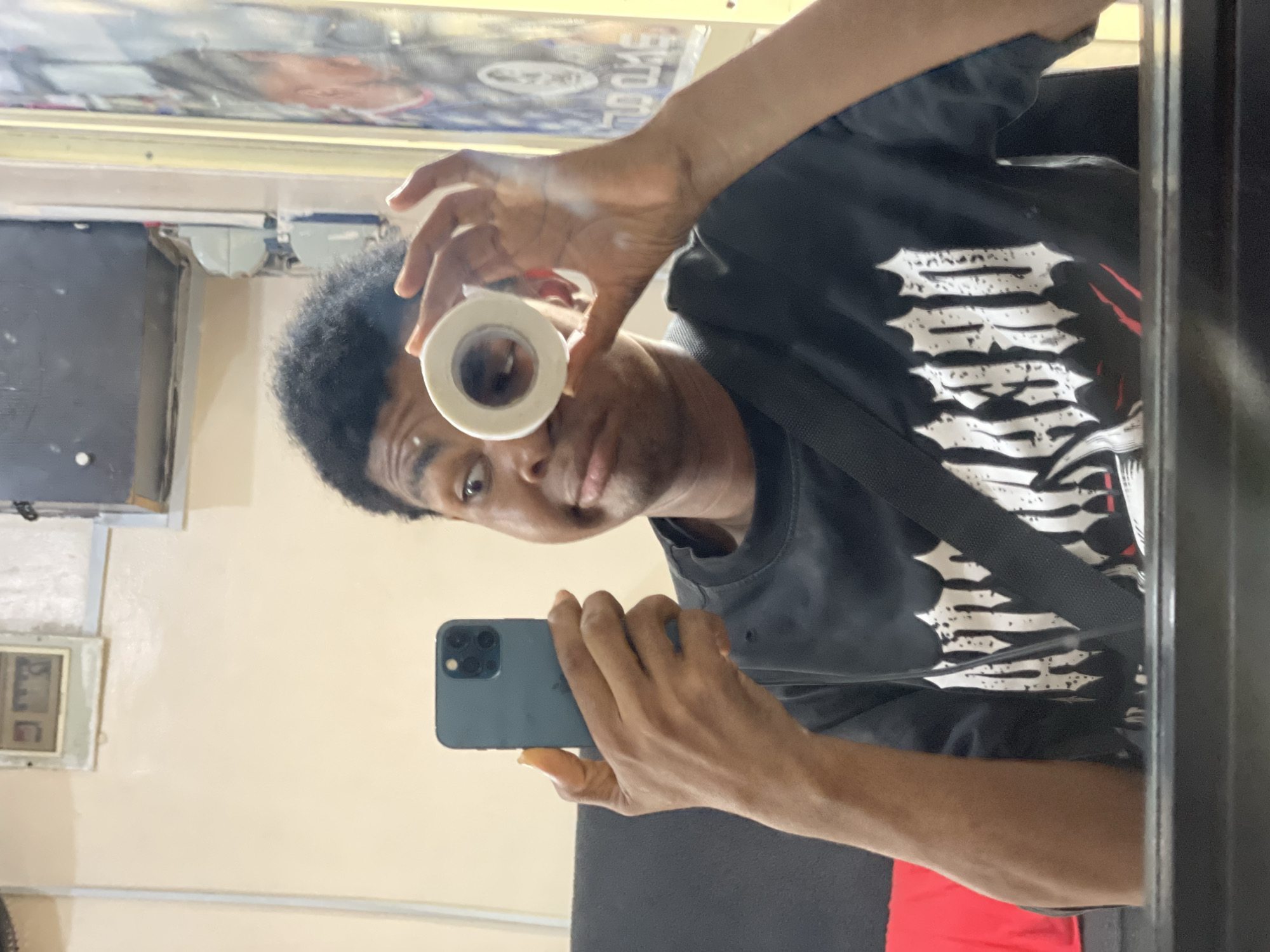 A man with dark, curly hair is taking a selfie in a mirror. He holds a roll of white tape to his left eye and a smartphone in his right hand. The man wears a black t-shirt with a white graphic design. A black strap crosses his chest. The reflection shows a light yellow wall with a framed object and electrical outlets. The location appears to be an interior space with a mirrored wall. The city of Ado, Nigeria is implied but not visually confirmed.
