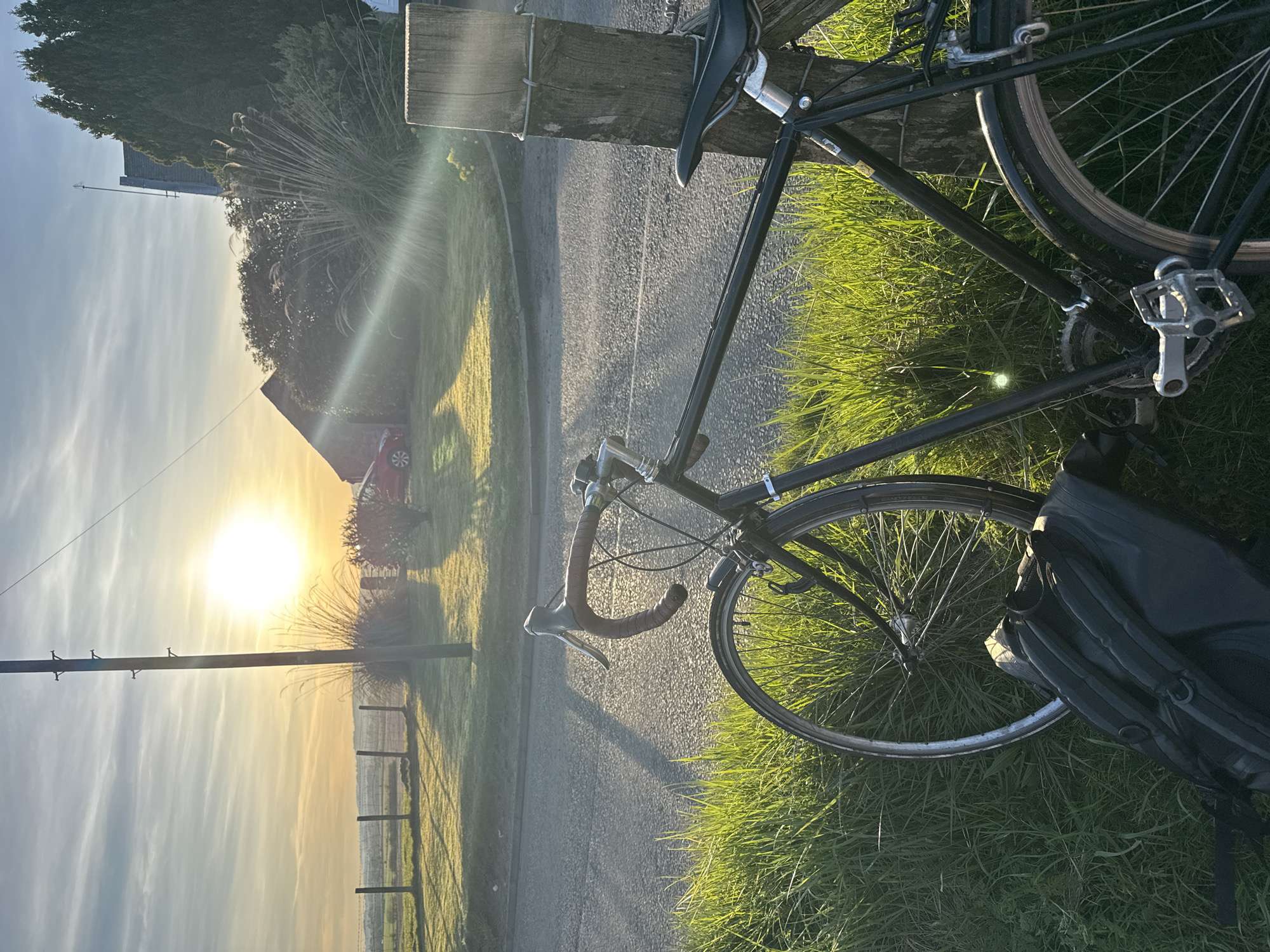 The image captures a serene, rural landscape during either sunrise or sunset, characterized by warm, golden light. A black bicycle is the main subject, leaning against a sturdy wooden post on the side of a narrow, possibly unpaved road or track.
The bicycle features drop handlebars with brown tape, a black saddle, silver pedals, and silver wheel rims. A black bag or pannier is attached to the rear of the bike, resting in the lush green grass of the verge. The grass is brightly illuminated by the low sun, creating a vibrant contrast with the darker bike frame and bag.
In the background, the sun is a bright, intense orb near the horizon, casting long shadows and a strong lens flare across the scene. A red car is partially visible near a building, which might be a house or barn. Further back, a utility pole stands tall, and a fence line stretches into the distance, suggesting expansive fields or open land beyond. The sky is clear with delicate, wispy clouds, bathed in the soft hues of dawn or dusk. The overall atmosphere is peaceful and calm, evoking a quiet moment in the countryside. No text is visible in the image.
