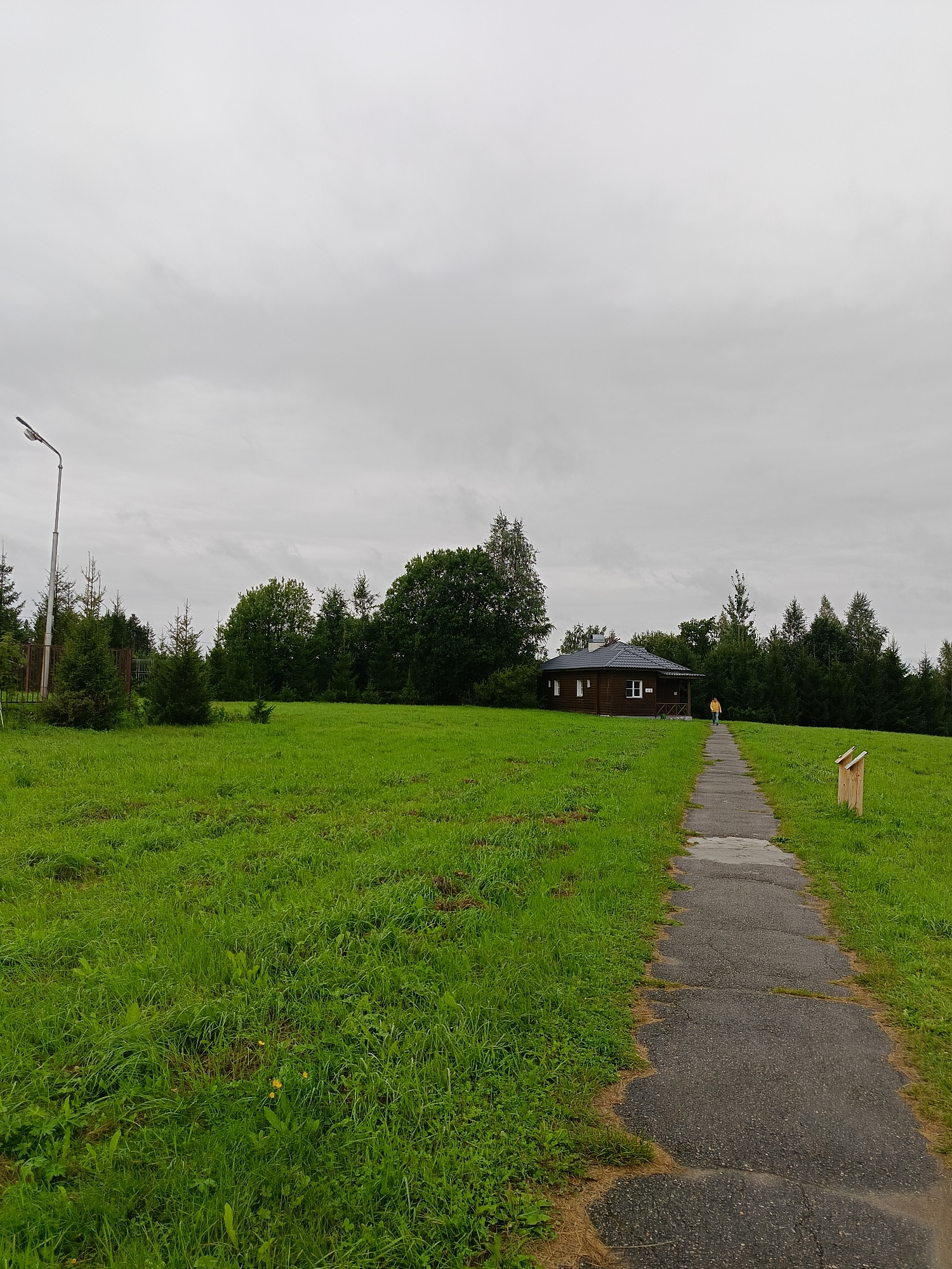 A paved walkway leads through a grassy field towards a small wooden house. The house has a dark roof, white-framed windows, and a dark-colored exterior. A person wearing a yellow jacket is walking away from the camera on the path towards the house. To the left of the path, a lamppost stands tall. The background is filled with a dense line of trees under a cloudy, overcast sky. A small wooden box is visible to the right of the path.