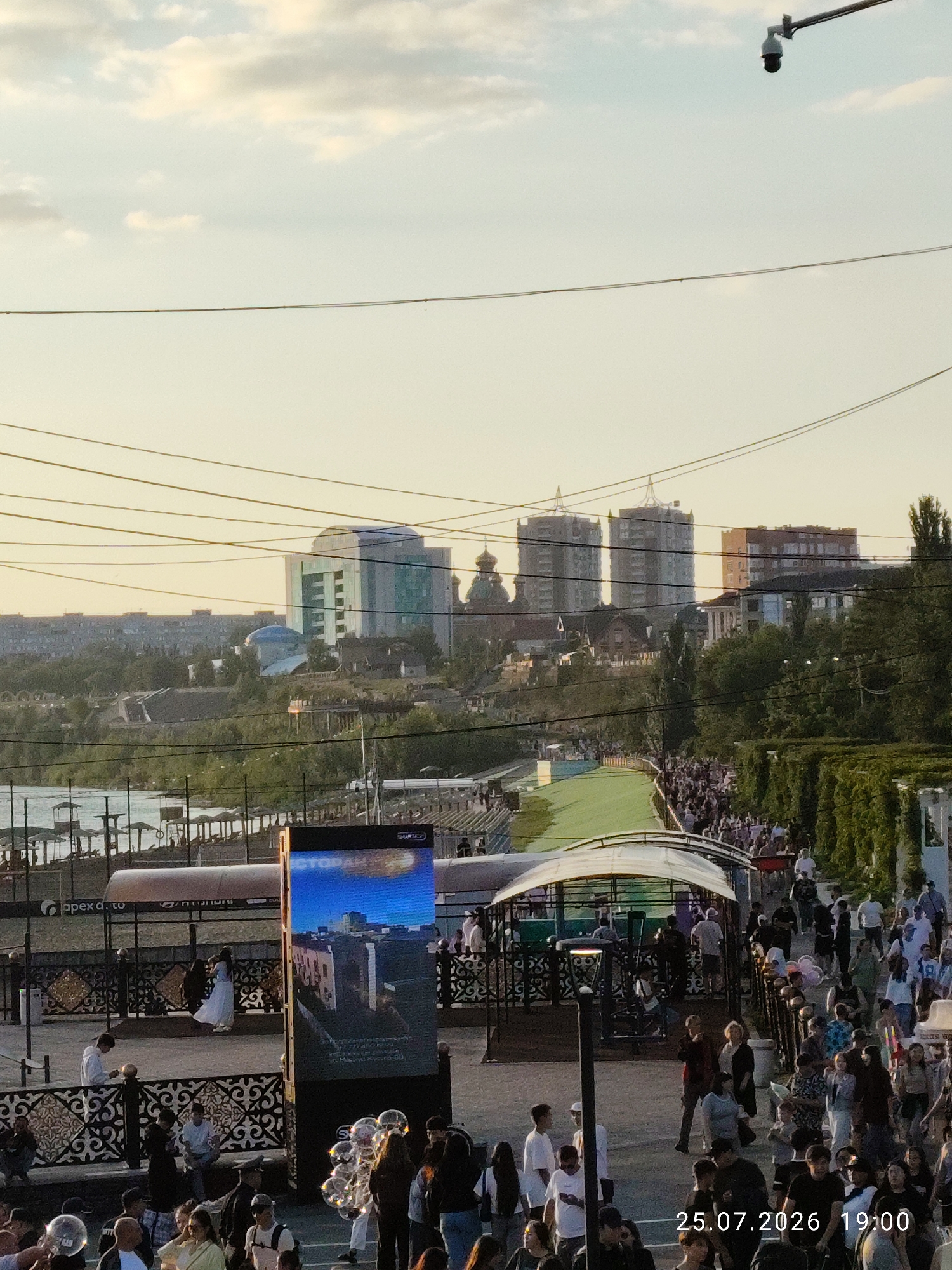 The image depicts a scene in Pavlodar, Kazakhstan, likely on the embankment or a waterfront area, given the presence of a body of water on the left. A large crowd of people is gathered, suggesting an event or a busy public space. The time stamp "25.07.2026 19:00" is visible in the lower right corner, indicating the date and time of the photograph.
In the foreground, a group of people are interacting, some are looking at a large LED screen which displays an image and text, possibly an advertisement or information. One person is holding a bunch of soap bubbles, which are floating in the air. Another individual is wearing a white dress and appears to be posing or walking. A man in a white shirt is seated on a bench, looking at his phone.
In the mid-ground, a paved walkway is lined with decorative metal fencing. Beyond this, a green, sloped area, possibly artificial turf, leads up towards a covered structure. More people are seen walking along this path. A line of trees and dense foliage forms a barrier on the right side of the image.
The background features a cityscape with various buildings, including modern high-rise structures and what appear to be older buildings with domes. Power lines crisscross the sky. A surveillance camera is mounted on a pole in the upper right portion of the image. The sky is partly cloudy with a soft, warm light, suggesting it is late afternoon or early evening. The presence of a body of water with structures resembling beach chairs or umbrellas on its bank further confirms a waterfront location.