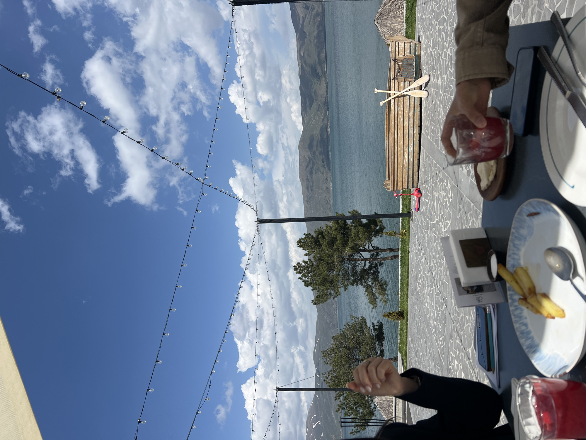 A laterally inverted image shows a table setting with partial views of two people at a restaurant overlooking a lake and mountains under a partly cloudy sky. The location is given as Unknown, Unknown.
In the foreground, a table is set with two plates of food, one with French fries and the other with a small appetizer, both partly eaten. Two drinks with red liquids, potentially juice or cocktails, are present. Silverware, napkins, and at least one book or pamphlet are also on the table. One person, wearing a dark jacket, has manicured nails and a ring visible on their hand. The other person, wearing a beige jacket, is holding one of the drinks.
In the background, beyond the table, there is a patio area constructed with patterned stone tiles. The patio leads to a wooden platform with two oars and a small, thatched-roof structure. The platform overlooks a large lake surrounded by mountainous terrain under a sky with scattered clouds. String lights are strung above the patio area.