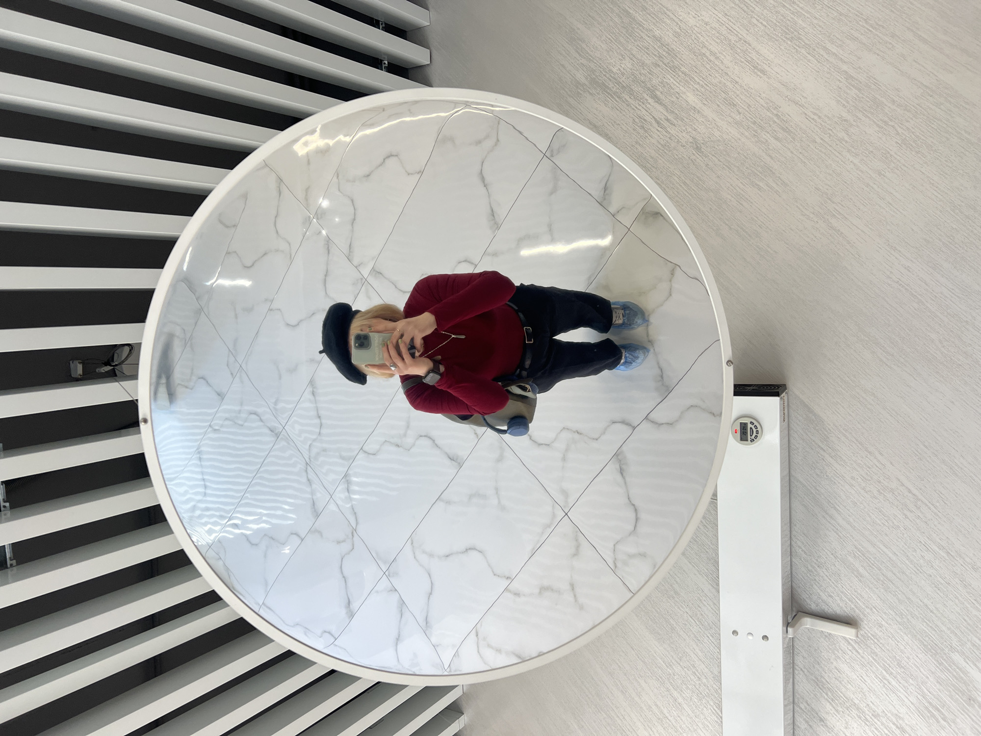 A person is taking a selfie in a round mirror. The reflection shows the person wearing a red sweater, black pants, and a black beret. The person is holding a smartphone in their hands. Their shoes appear to be covered in blue plastic bags. The mirror is placed on a stand. The background includes a wall with black and white stripes and another wall with a textured light gray finish. A white object with a digital display is attached to the wall next to the mirror. The location appears to be indoors. Odesa, Ukraine, is not discernible from the image.