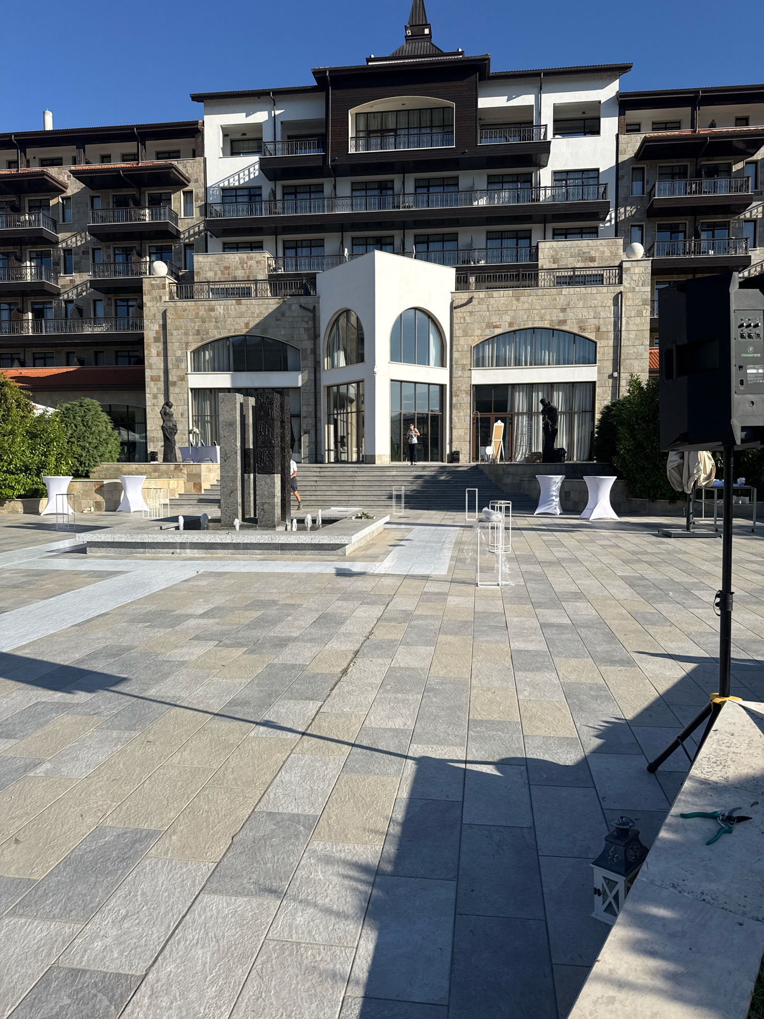The image depicts the exterior of a hotel or resort in Razliv, Bulgaria. The building is large and multi-story, with a facade of stone and white plaster. There are many balconies with dark railings. In front of the building is a large paved courtyard with a fountain in the center. Several white-covered cocktail tables are set up in the courtyard, along with some white decorative stands and a dark speaker on a stand. A person is visible near the entrance of the building. The sky is clear and blue.