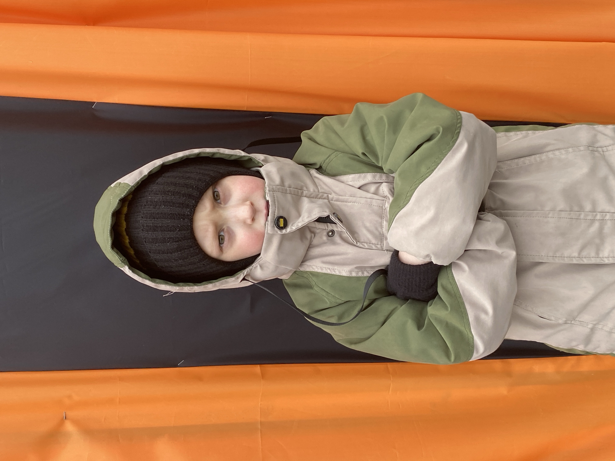 A young child wearing a black knit cap and a beige and green winter coat is lying on a black surface, which is in turn on top of an orange surface. The child has their arms crossed. The location is not discernible from the image.