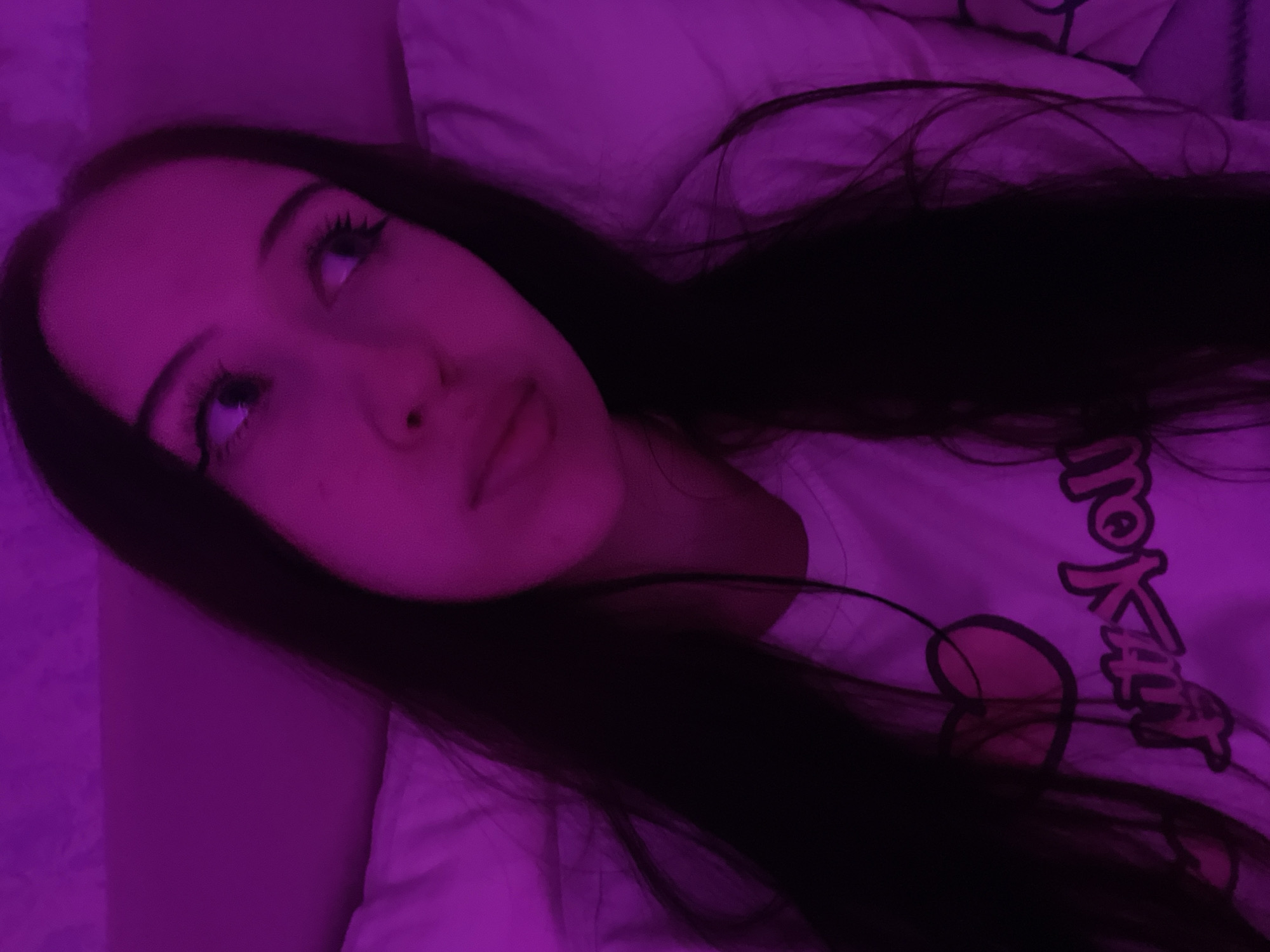 This is a close-up, low-angle selfie of a young woman with long, dark hair, lying down and looking upwards. She is illuminated by purple lighting, which casts a vibrant glow on her face and the white bedding she is on. Her eyes are open, and her gaze is directed away from the camera, giving her a thoughtful or perhaps dreamy expression. Her makeup includes pronounced eyeliner and eyelashes. She is wearing a white t-shirt with what appears to be a cartoonish graphic and text printed on it. The visible text appears to be Japanese characters, possibly part of a brand or design. The overall mood is intimate and serene, possibly taken at night or in a dimly lit room given the artificial lighting. There are no discernible cues in the image that would specifically place the location as Votkinsk, Russia, beyond what is provided in the prompt.