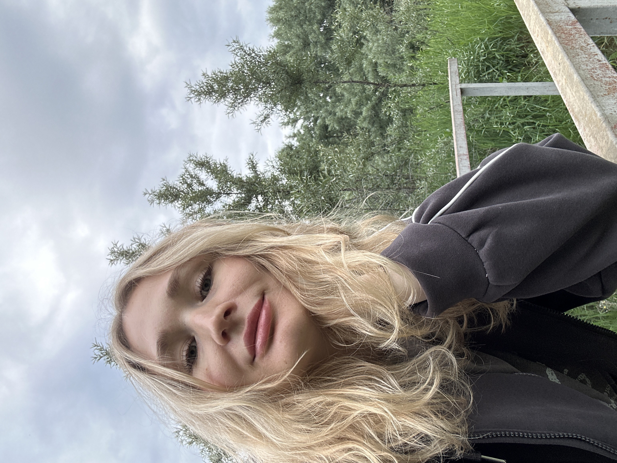 A young Caucasian woman with blonde, wavy hair is taking a selfie outdoors. She is wearing a dark gray zip-up hoodie with white piping down the sleeves. Her right hand is resting on her cheek, and her left arm is partially visible. The background consists of lush green foliage, including trees and tall grass. In the foreground, a weathered metal railing is visible, suggesting the woman might be on a bridge or a raised walkway. The sky is overcast with gray clouds. The location is not specified in the image or context.