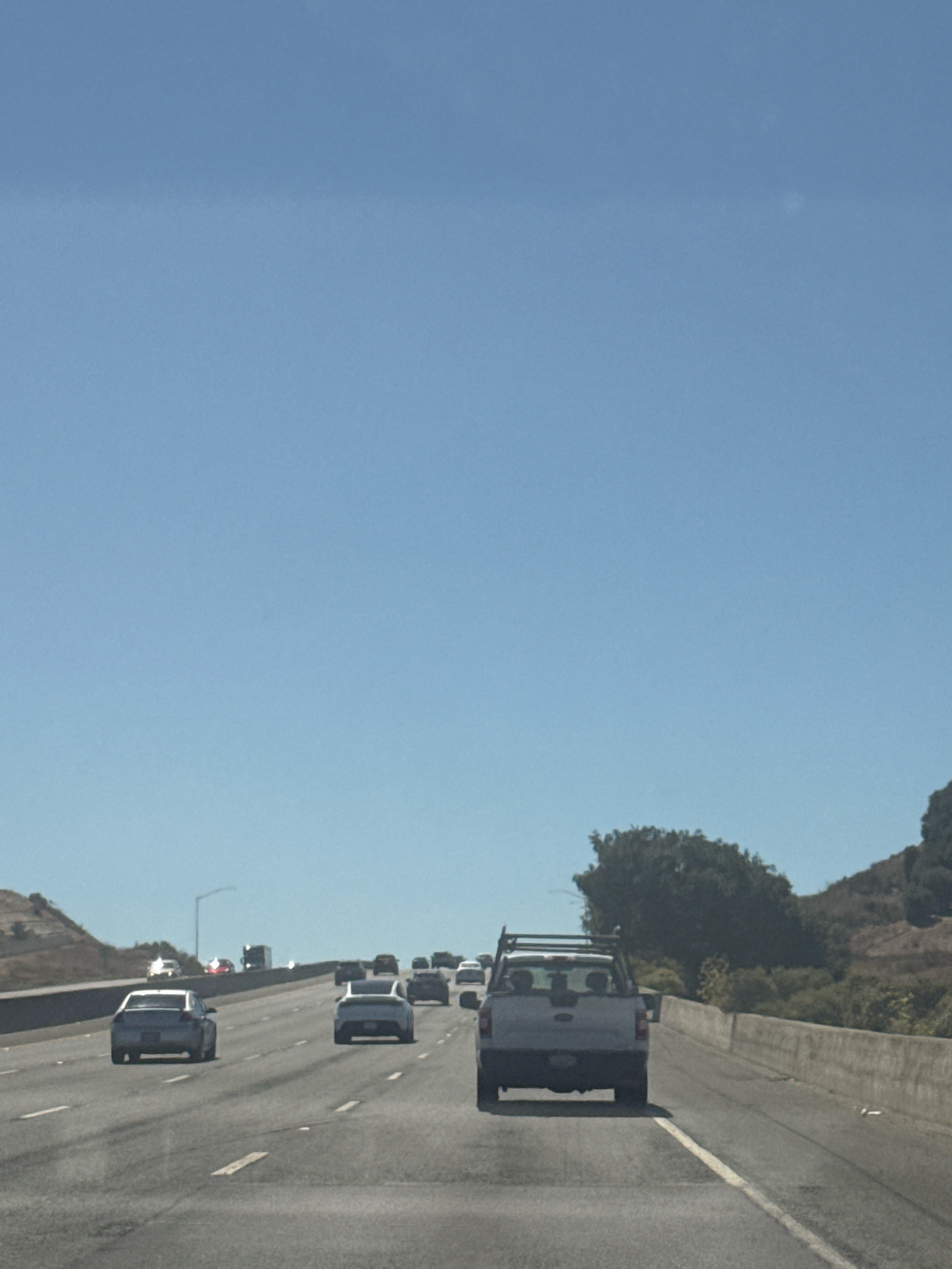 A daylight, eye-level, medium shot captures a freeway scene with traffic moving under a clear blue sky. The road is asphalt with white lane markings and appears to be part of a highway with retaining walls on the right. The image is focused on the vehicles, with the foreground occupied by a light-colored pickup truck with a rack on top. Several cars are visible in front of and behind the pickup, traveling in the same direction. In the distance, more vehicles and trees lining the sides of the freeway can be seen. The overall environment suggests a sunny day on a highway, possibly in or near American Canyon, California, given the vegetation and terrain that resembles the region.