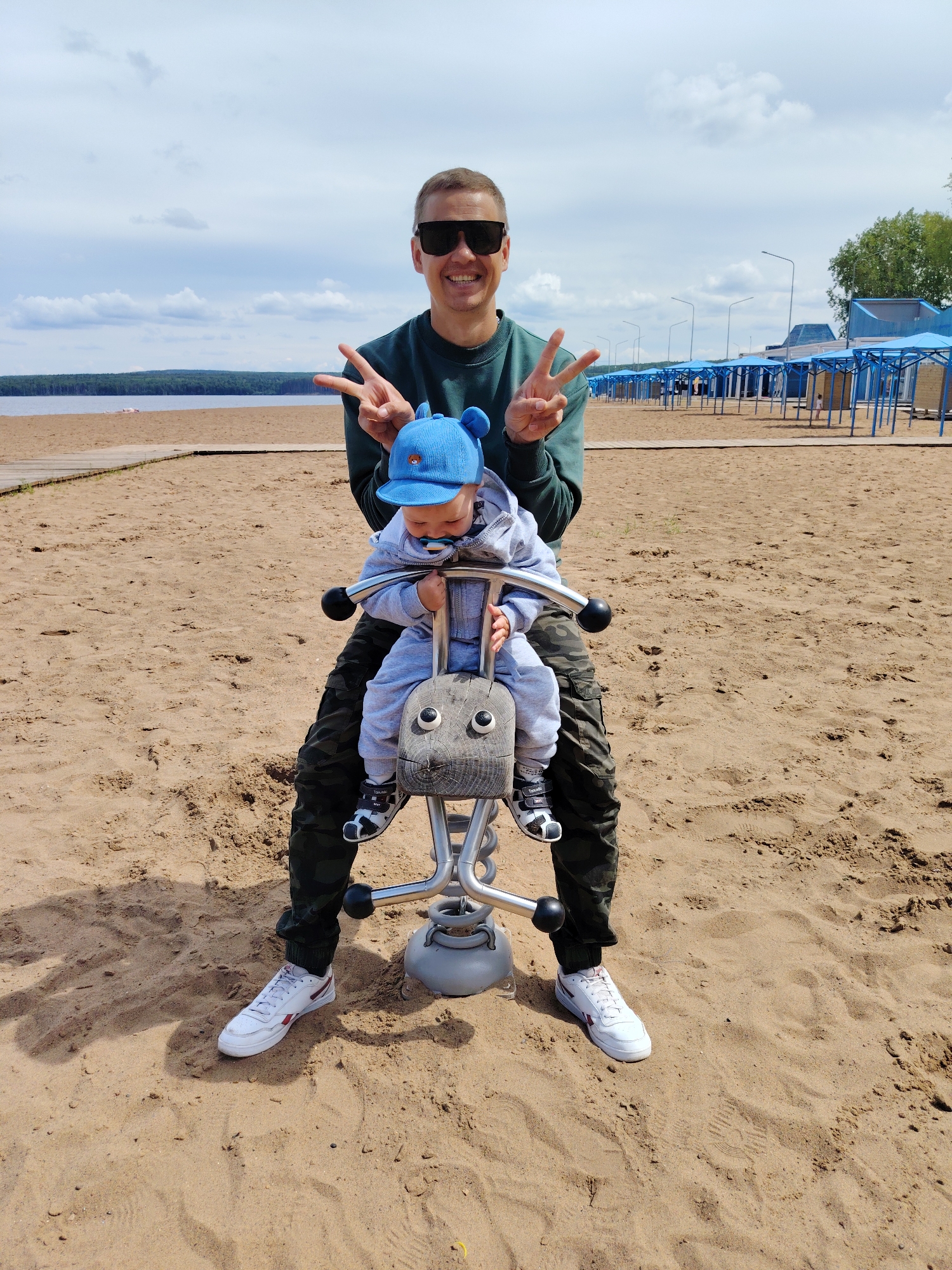 The image depicts a man and a baby/toddler enjoying a sunny but cloudy day on a sandy beach in Chaikovsky, Russia. The man, positioned behind a spring rider, is wearing sunglasses, a dark green long-sleeved shirt, camouflage pants, and white sneakers with red accents. He is smiling brightly at the camera and making peace signs with both hands.
Seated on the spring rider is a baby or toddler, dressed in a light grey hooded jumpsuit, a vibrant blue hat with ear-like protrusions, and black and white sandals. A pacifier is visible in the child's mouth. The spring rider itself has a wooden animal-shaped head with painted eyes, mounted on a metallic frame with a grey base.
The setting is a wide, sandy beach, with a wooden boardwalk running along the left side of the frame. In the background, a large body of water, likely a lake or wide river, stretches towards a distant treeline under a cloudy sky. To the right, a row of blue-roofed cabanas or changing huts and tall streetlights line the beach, indicating a developed recreation area. The overall mood is cheerful, emphasized by the man's happy expression. Visible text includes "TOMMY" on the strap of the baby's sandal.