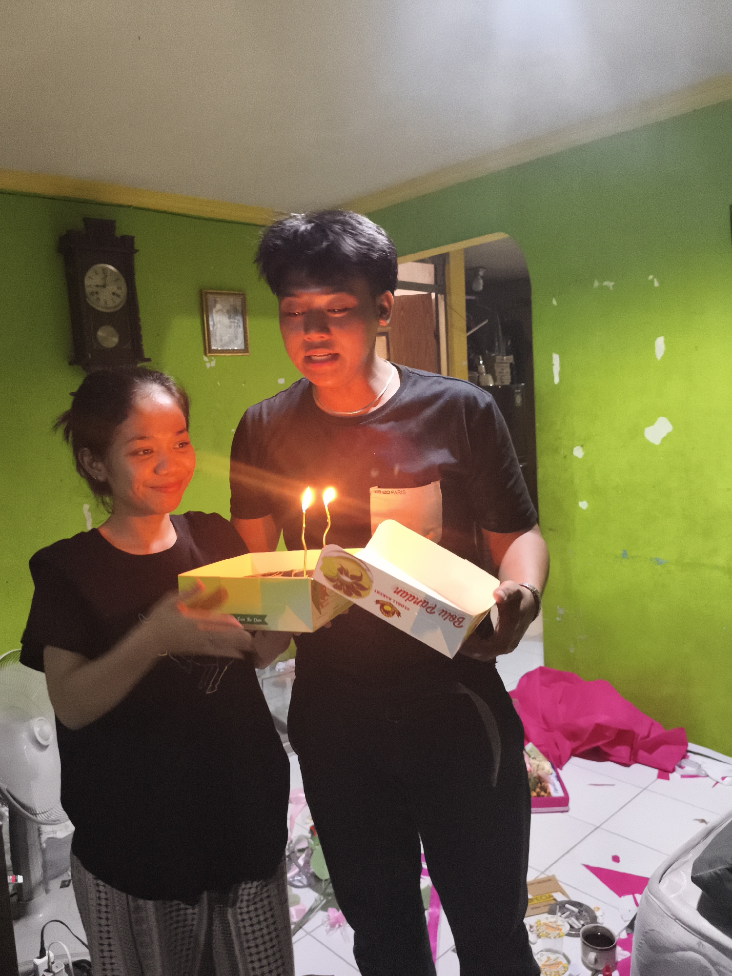 Two young adults, a woman and a man, are standing in a room with lime green walls. The woman, on the left, is wearing a black t-shirt and patterned pants. She is holding a yellow cake box with two lit candles and is smiling. The man, on the right, is wearing a black t-shirt and black pants. He is holding a white cake box and looking at the woman. In the background, there is a large antique clock on the wall and a framed picture. A white oscillating fan is visible on the left. The floor is tiled and there are some pink pieces of paper scattered on it. A pile of pink fabric is on the floor to the right.
The scene appears to be a birthday celebration. The overall mood is joyful and celebratory, as indicated by the smiles of the subjects and the presence of a birthday cake with candles. The lighting suggests it might be evening or indoors with artificial lighting. The text visible on the cake box held by the man reads "Bolu Pandan" and "GLOBAL BAKERY". The text on the pocket of the man's shirt is partially visible and appears to say "KENZO PARIS". The location context is Depok, Indonesia.