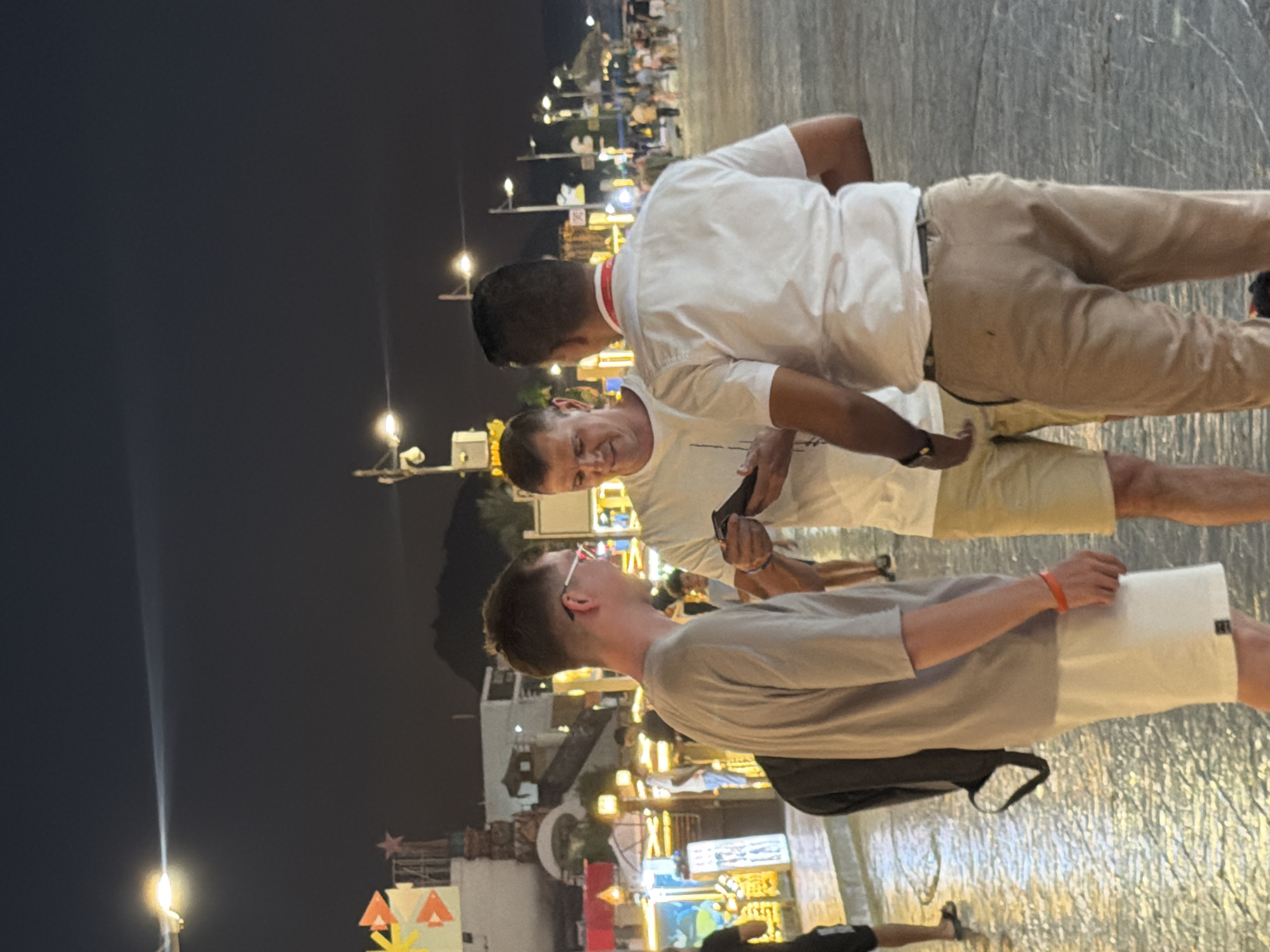The image shows three men standing on a paved street at night. The man on the left is wearing a grey t-shirt and white shorts, with glasses and a red wristband. The man in the middle is wearing a white t-shirt and beige shorts, and is holding a phone. The man on the right is wearing a white short-sleeved shirt with a red lanyard and beige pants. They appear to be engaged in a conversation. The background shows a street scene with buildings, lights, and other people walking. The lighting suggests it is a commercial area, possibly a tourist district. The presence of streetlights and illuminated signs indicate an urban environment. The general atmosphere is that of a lively evening in a city. Without further context, the specific location within Sharm El Sheikh, Egypt, cannot be definitively identified, but it resembles a promenade or a busy street with shops and entertainment venues.