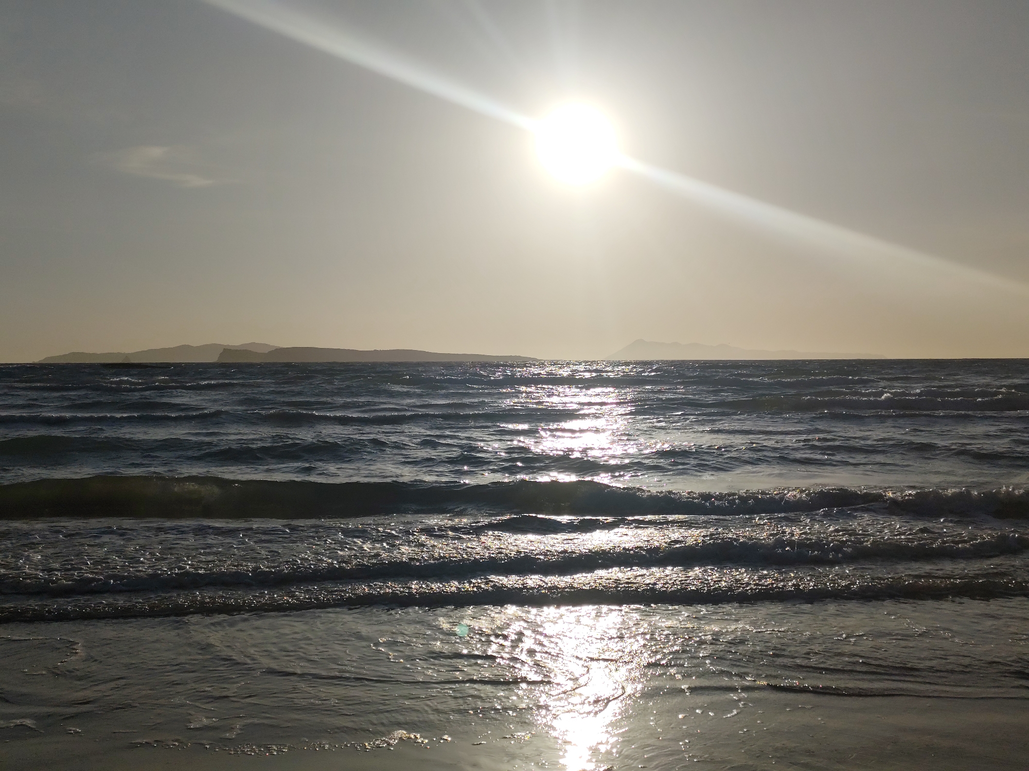 The image depicts a sunlit seascape, likely captured in the coastal area of Agios Stefanos, Greece. The sun is positioned high in the sky, casting a bright glare and creating a prominent reflection on the water's surface. The sea is moderately choppy, with small waves visible closer to the shore. The water reflects the sunlight, creating a shimmering effect. In the distance, a faint outline of land is visible, likely a neighboring island or a distant part of the coast. The sky appears hazy or overcast, contributing to the overall brightness of the scene. No people or distinct objects are visible in the image apart from the natural elements. The overall atmosphere suggests either early morning or late afternoon light conditions.