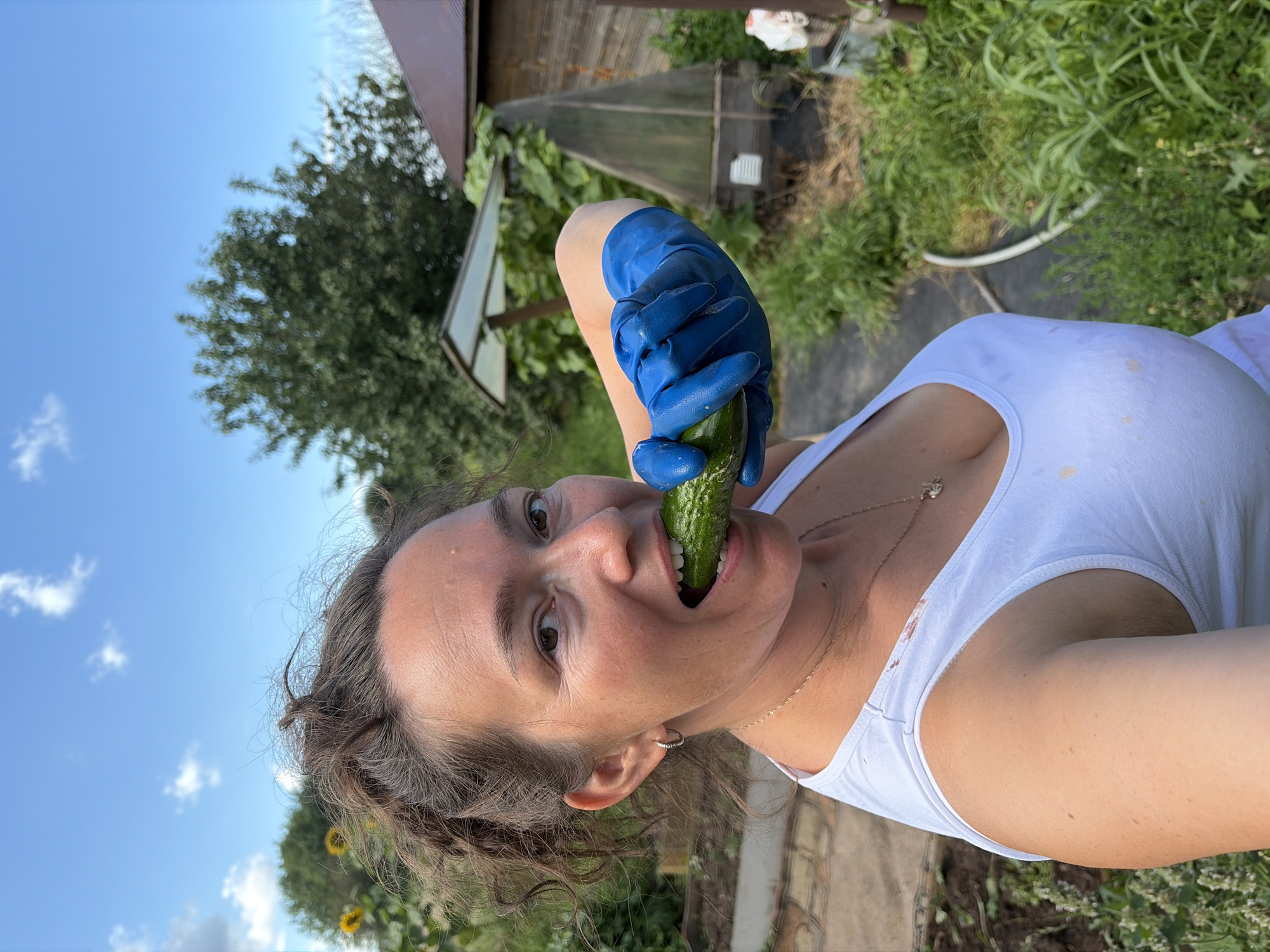 A woman wearing blue rubber gloves and a white tank top is biting into a cucumber. She has brown curly hair and is looking at the camera. The scene appears to be outdoors in a garden, with trees and a wooden structure in the background. The sky is blue with some clouds. The location is identified as Olkhovka, Russia.