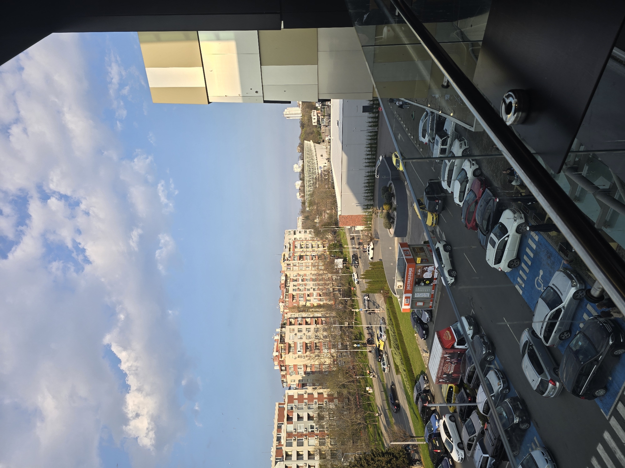 The image depicts an elevated view of a street scene in Burgas, Bulgaria, likely taken from a high-rise building. The sky is partially cloudy with patches of blue. The street below is lined with parked cars, including several in marked handicapped spaces. There is a small shop stand on the side of the road that sells insurances. Residential buildings of varying heights and color palettes are seen along the opposite side of the street. Distant structures are also visible, including what looks like a stadium or sports complex.