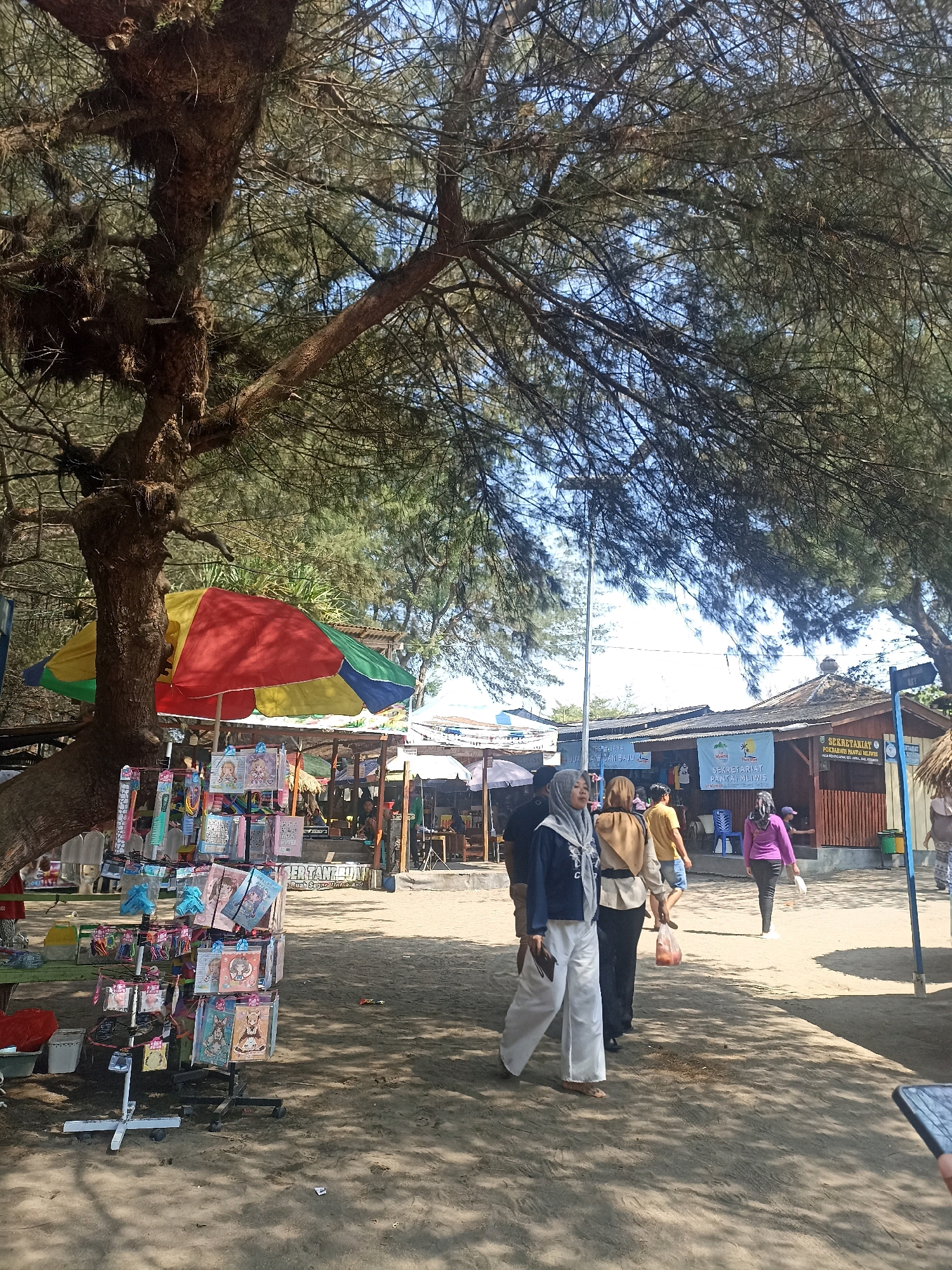 In Kenoyajaya, Indonesia, a sunny day at a beachside market is depicted. The foreground features a display rack filled with various merchandise, including anime-themed items and accessories. A large, weathered tree with sparse foliage partially obscures the sky and casts dappled shadows across the sandy ground. To the left of the tree, a colorful, multi-paneled umbrella stands open, shades a small stall.
Several people are walking and browsing in the midground. Two women, one in a grey hijab and white pants, the other in a beige headscarf and black pants, are walking together. A man in a yellow t-shirt walks away from the camera, and another woman in a purple top and black pants is seen further in the background. Several small stalls or shops with thatched roofs are visible behind the shoppers, some displaying signage. One sign reads "SEKRETARIAT POLICHARISTUS PANTAI MELATI." The overall environment is sandy, suggesting a beach location.