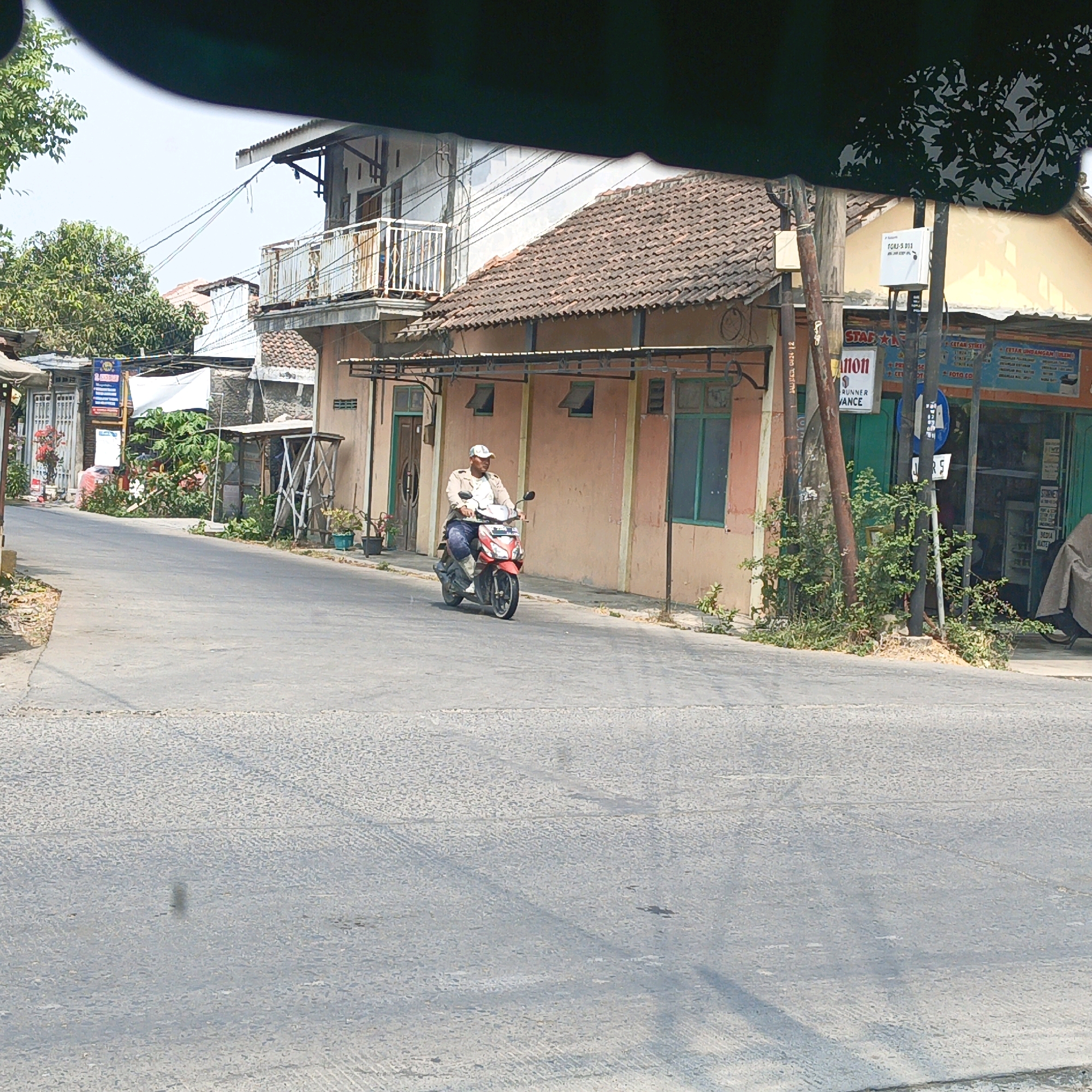 A man is riding a red motorcycle down a street in Pegandon, Indonesia. The man is wearing a white baseball cap, a light-colored jacket, and dark pants. He is riding on the left side of the road. The street is paved and appears to be in good condition. There are buildings on either side of the street, as well as trees and plants. The buildings are a mix of residential and commercial properties. The sky is clear and blue.
