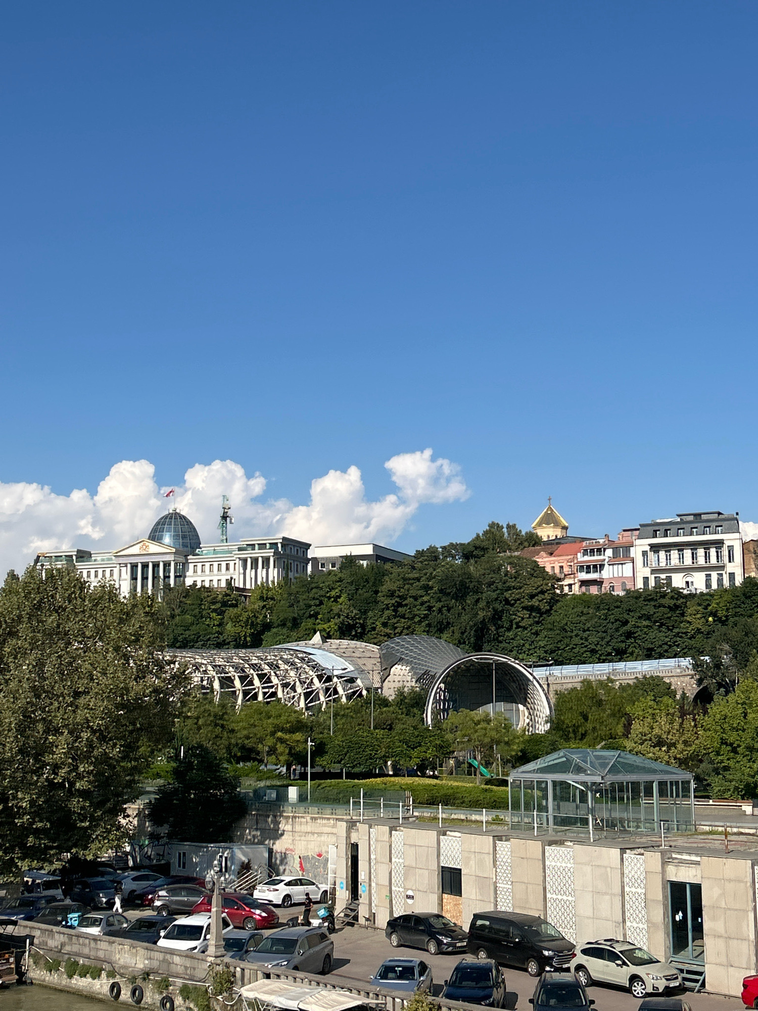 This is a wide shot of a city skyline, likely in Tbilisi, Georgia, under a clear blue sky with scattered white clouds. The most prominent structure in the upper half of the image is a large white building with a dome, possibly a government building or palace, situated on a hill surrounded by lush green trees. In front of this building, on the slope of the hill, is a modern architectural structure with a curved, metallic roof. Below this, a parking lot is visible, filled with various cars. Further down, a glass-enclosed structure is situated on a concrete platform. The image also shows a body of water in the lower left corner, suggesting it might be a river.