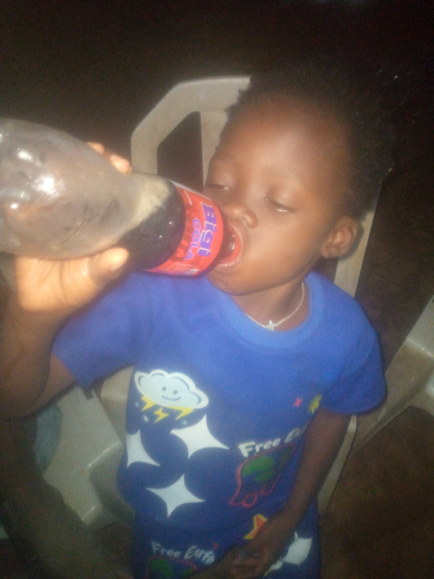 A child is drinking from a large plastic bottle of "Bigi Cola" while seated in a white plastic chair. The child is wearing a blue shirt with a cartoon cloud, lightning bolts, and stars on it. The background is dark and indistinct. The city and location are unknown.