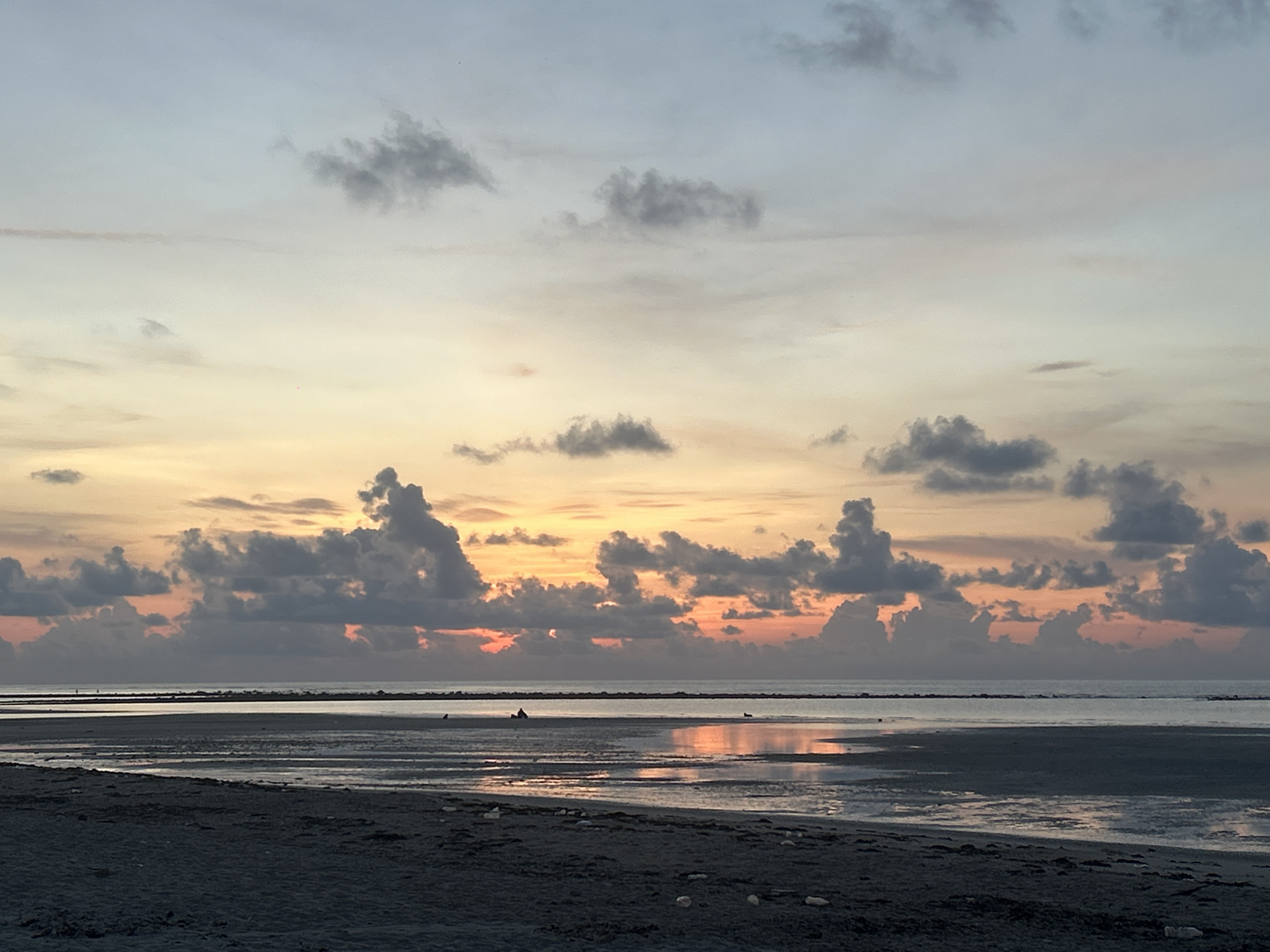 The image depicts a coastal scene at dusk or dawn, likely in Baan Harn, Thailand. The foreground is a dark sand beach with visible water reflecting the sky. Further out, the water extends to a visible breakwater or submerged barrier in the midground. Clouds are scattered across the sky, with warm colors of orange and yellow peeking through the cloud cover near the horizon. A single, small figure is visible in the wet sand area. Debris is scattered across the sand.