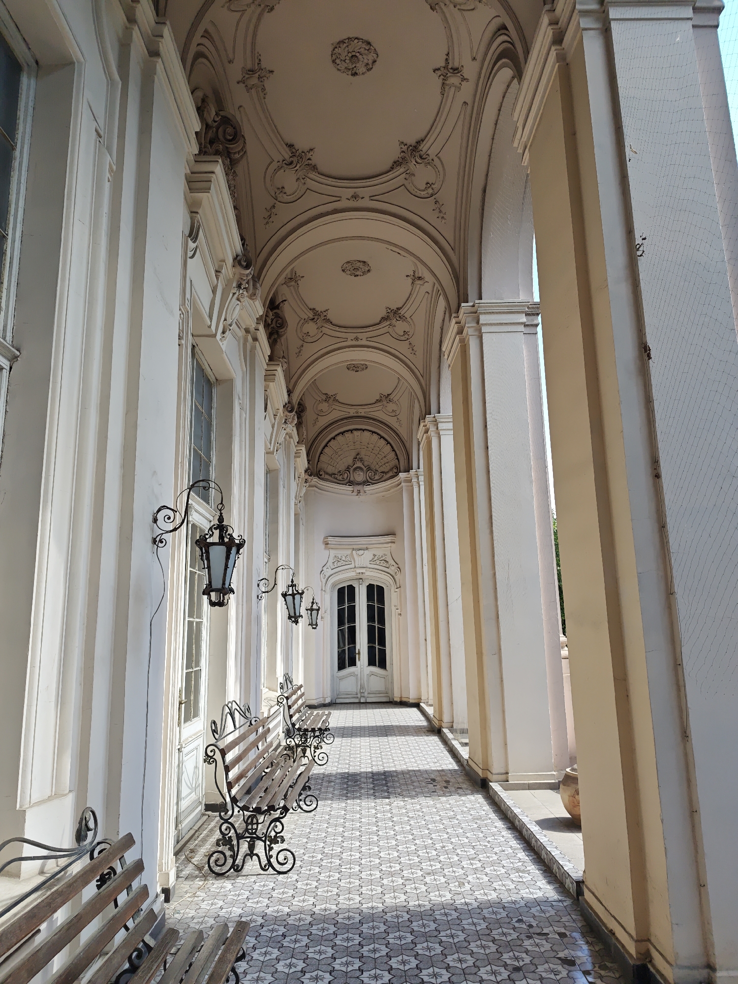 The image depicts a covered walkway or arcade with a tiled floor and arched ceiling, located in the city of Lviv, Ukraine. The architecture features ornate detailing on the ceiling and elegant white columns. Several wooden benches with decorative wrought-iron bases are placed along the walkway, suggesting a public space for rest. Hanging lanterns with dark metal frames and frosted glass shades are mounted on the wall, adding to the ambiance. The walkway leads towards a set of double doors with glass panels at the far end, hinting at an entrance to a building. Sunlight casts shadows on the tiled floor, indicating a bright day. The overall impression is one of historical grandeur and a tranquil outdoor setting.