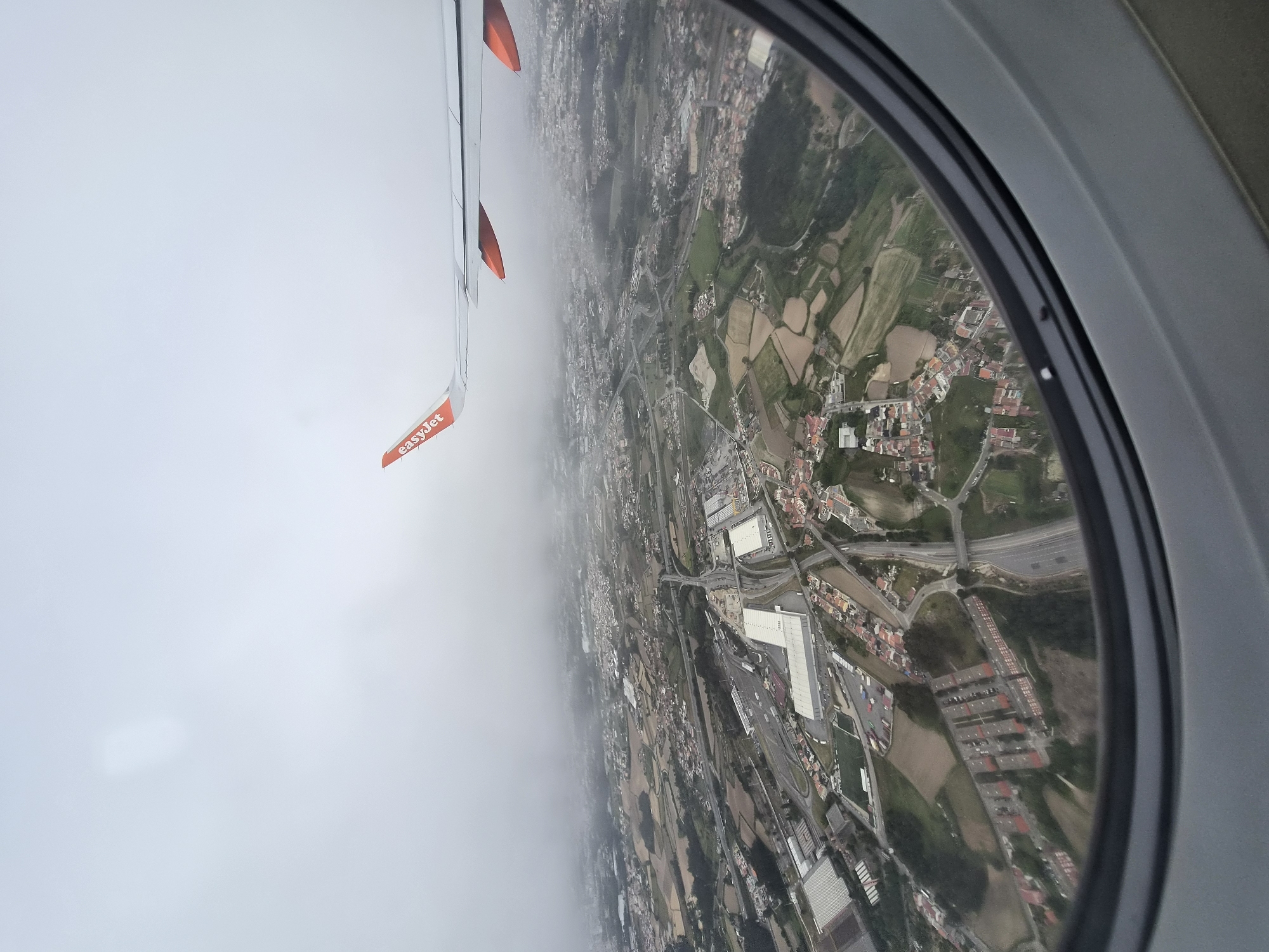 The image shows a view from an EasyJet airplane window, looking down over Matosinhos, Portugal. The wing of the plane is visible in the upper left, with an orange "easyJet" flag attached. The view below reveals a landscape comprised of a mix of urban and rural areas. There are densely populated residential areas, industrial zones with large warehouse-type buildings, fields, and roads including highway overpasses. The weather appears overcast, with some areas obscured by fog or low clouds.