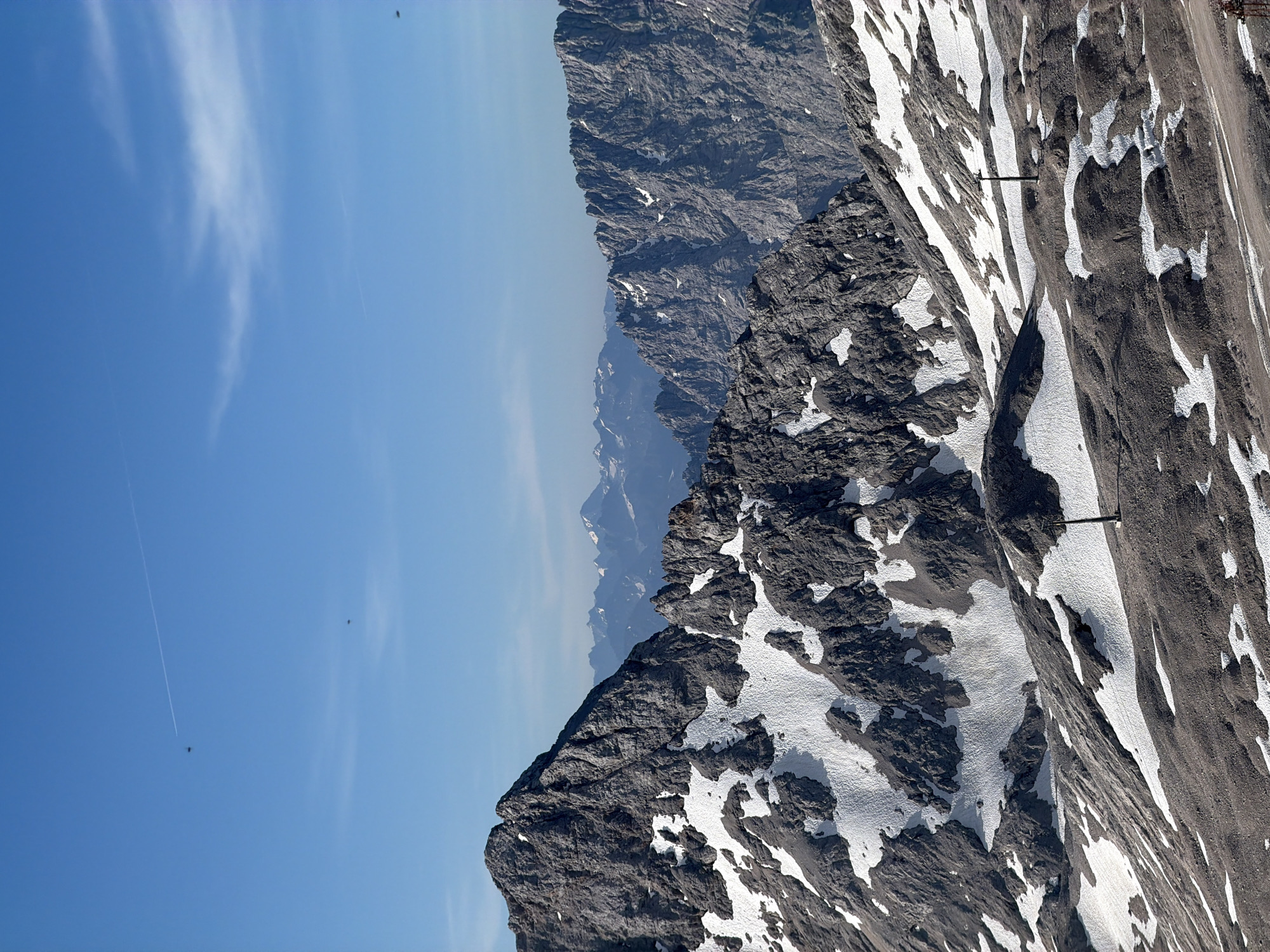 The image depicts a panoramic view of a mountainous landscape, likely at a high altitude, given the presence of snow patches on dark, rocky terrain. The sky is a clear, bright blue with a few wispy clouds and two faint contrails from aircraft. In the foreground, the rugged, grey, rocky peaks are partially covered with white snow. The terrain slopes downwards, revealing more rocky ground interspersed with snow. There are some dark, slender poles visible on the slopes, possibly part of a ski lift or other mountain infrastructure. In the mid-ground and background, a range of further, more distant, jagged mountain peaks can be seen, some also dusted with snow. The overall impression is one of a vast, natural mountain environment under a clear sky. This scene is consistent with the views from the Zugspitze, Germany's highest peak, located near Garmisch-Partenkirchen.