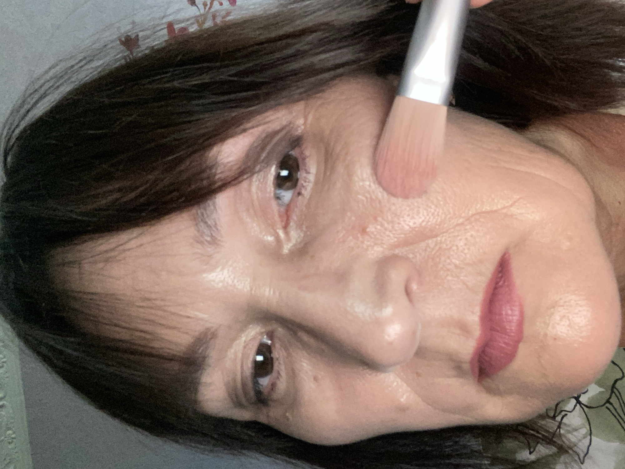A close-up, head-and-shoulders shot shows a woman with dark brown hair and bangs applying blush to her right cheek with a pink-bristled brush. Her eyes are open and looking towards the camera. She is wearing lipstick. The background is a neutral, textured wall. There is no visible indication of the location being Novokuznetsk, Russia.