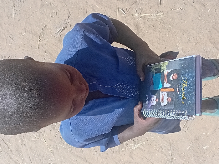 A high-angle shot depicts a young child, appearing to be male, standing outdoors on a light brown, unpaved sandy surface with scattered dry vegetation in Ayama, Nigeria. The child wears a blue short-sleeved top with white embroidered patterns on the chest and sleeves, light-colored trousers, and light-colored footwear. The child holds a spiral-bound notebook with both hands, looking down at its cover. The notebook cover is dark, featuring four small portrait photographs arranged in a grid, with visible text including "Thanks" in a decorative font and "J. Godwin." The child's head is tilted forward, obscuring most of their face as they focus on the notebook. The scene is illuminated by natural daylight.