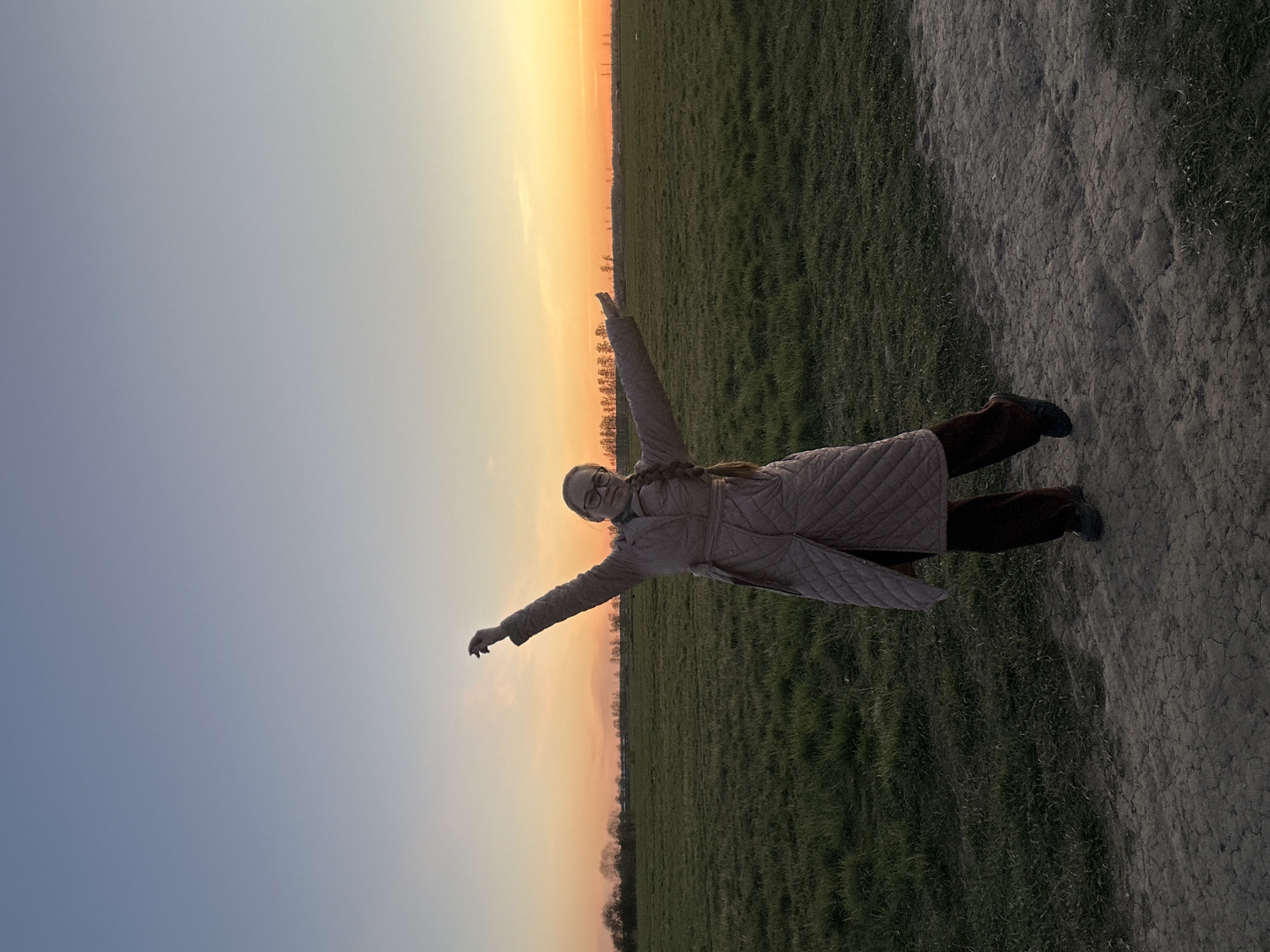 A woman in a pink coat, glasses, and dark pants poses on the ground with her arms outstretched against a sunset sky. She is located in a grassy field next to a dirt path. The sun is setting on the horizon. The city of Saint Petersburg, Russia, is not evident in this image.