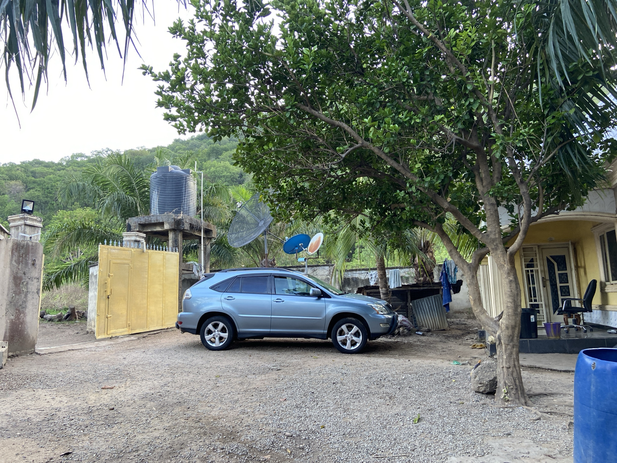 The scene is a residential area in Mararaba, Nigeria. A silver SUV is parked on a gravel driveway next to a yellow gate. In the background are trees, a water tower, and satellite dishes. A building with a yellow exterior is visible to the right of the frame. Laundry hangs outside near the building. A blue plastic barrel is visible in the foreground, and a large tree partly obscures the building.
