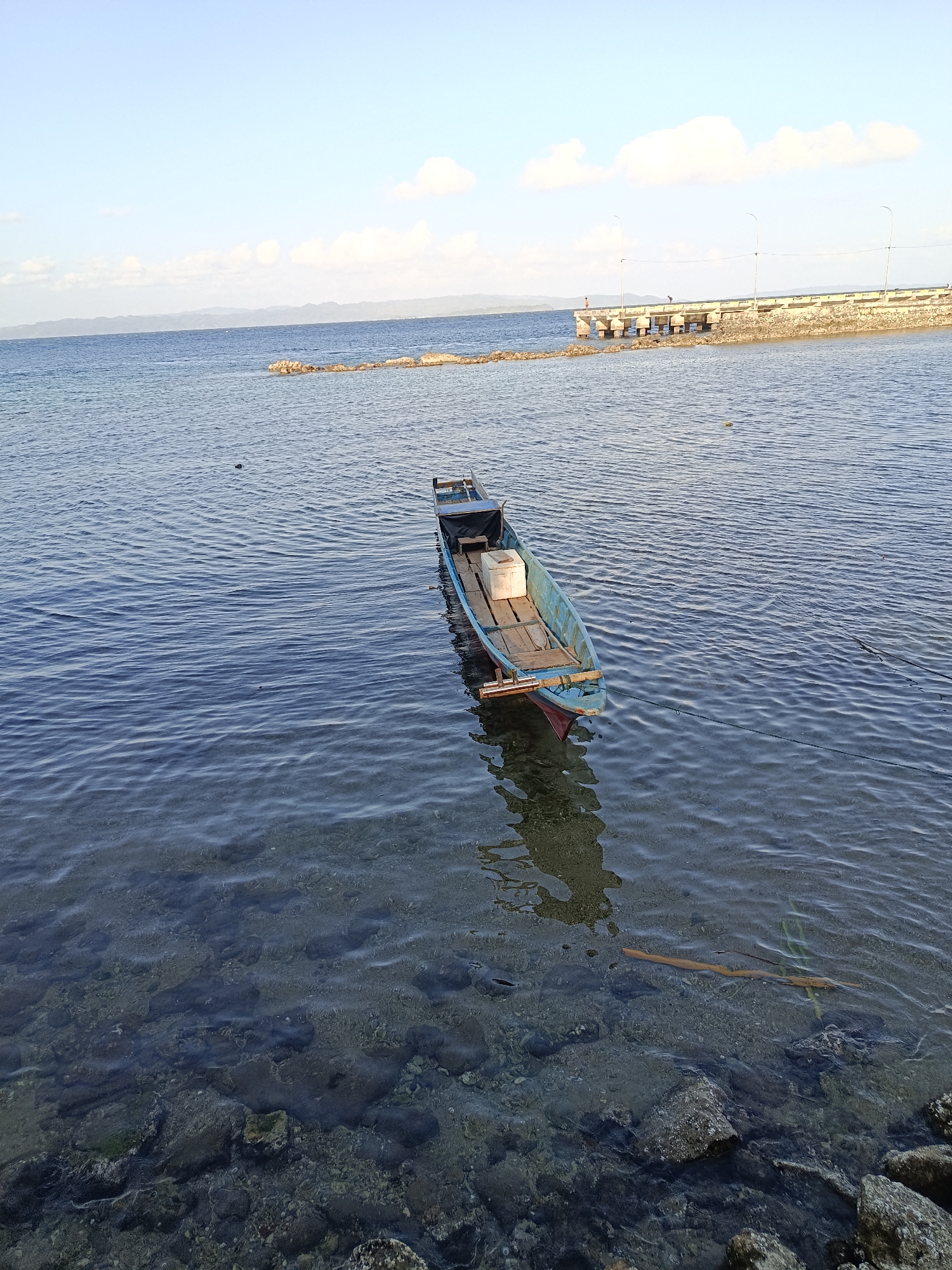 In Mansalean, Indonesia, a small, light blue wooden boat is anchored in clear, shallow water. The boat, which appears to be a fishing boat, contains a white cooler and some wooden planks. The seabed is visible through the water, littered with rocks and small marine life. In the distance, a concrete pier extends into the water, lined with lampposts. The sky is a pale blue with scattered white clouds, and hazy mountains are visible on the horizon. The overall scene is tranquil, capturing a peaceful moment by the sea.