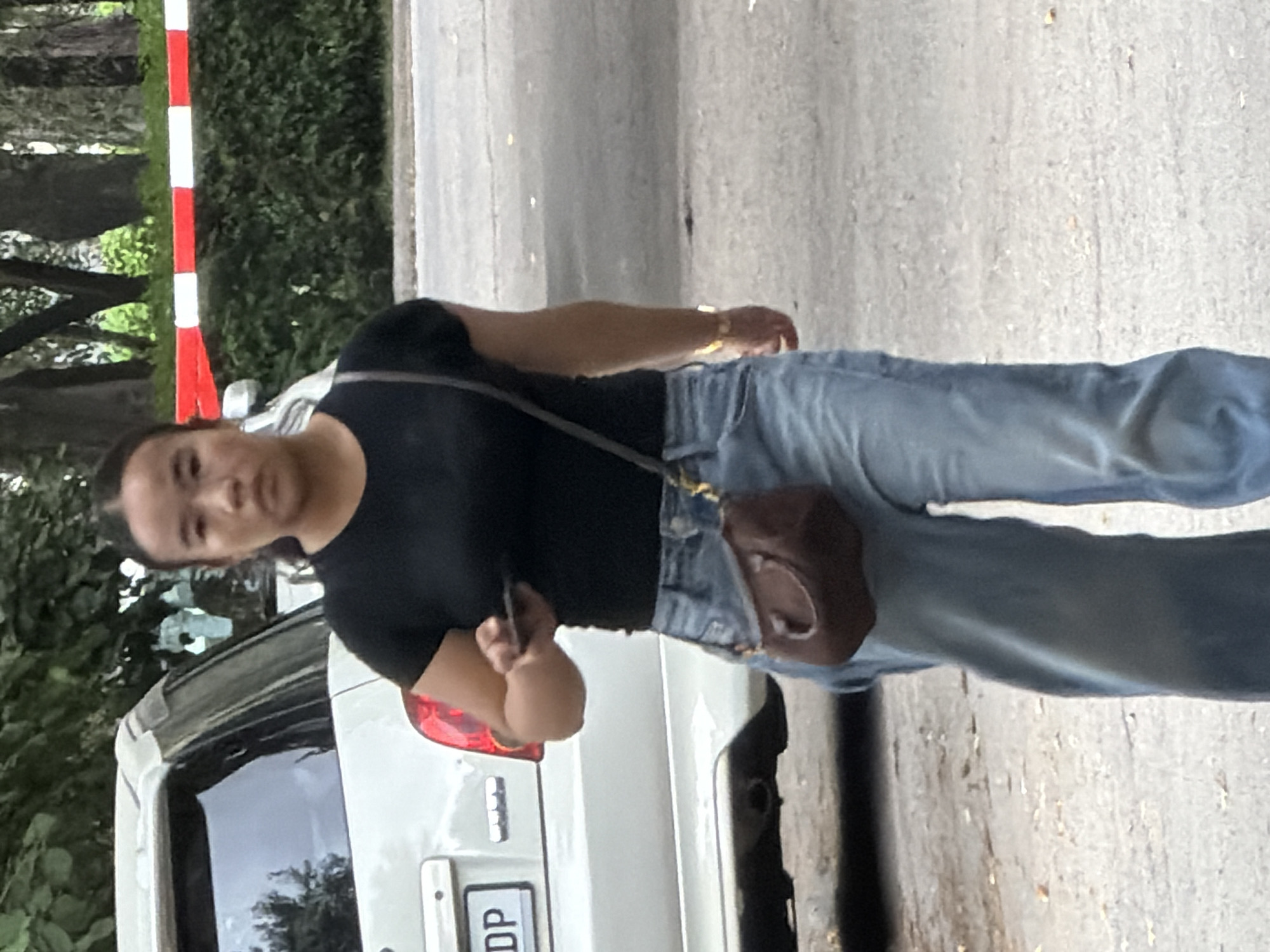 A woman wearing a black t-shirt and blue jeans stands next to a white car. She has her hair pulled back and is looking at her phone, which she holds in her right hand. A small brown purse is slung across her body. The car has a license plate with the letters "DP" visible. Behind the woman and to the left is a red and white striped pole and dense green foliage. The ground is a rough, gray surface, possibly asphalt or concrete. The lighting suggests it is daytime, and the scene appears to be outdoors, possibly on a street or in a parking area. The woman's expression is neutral as she focuses on her phone.