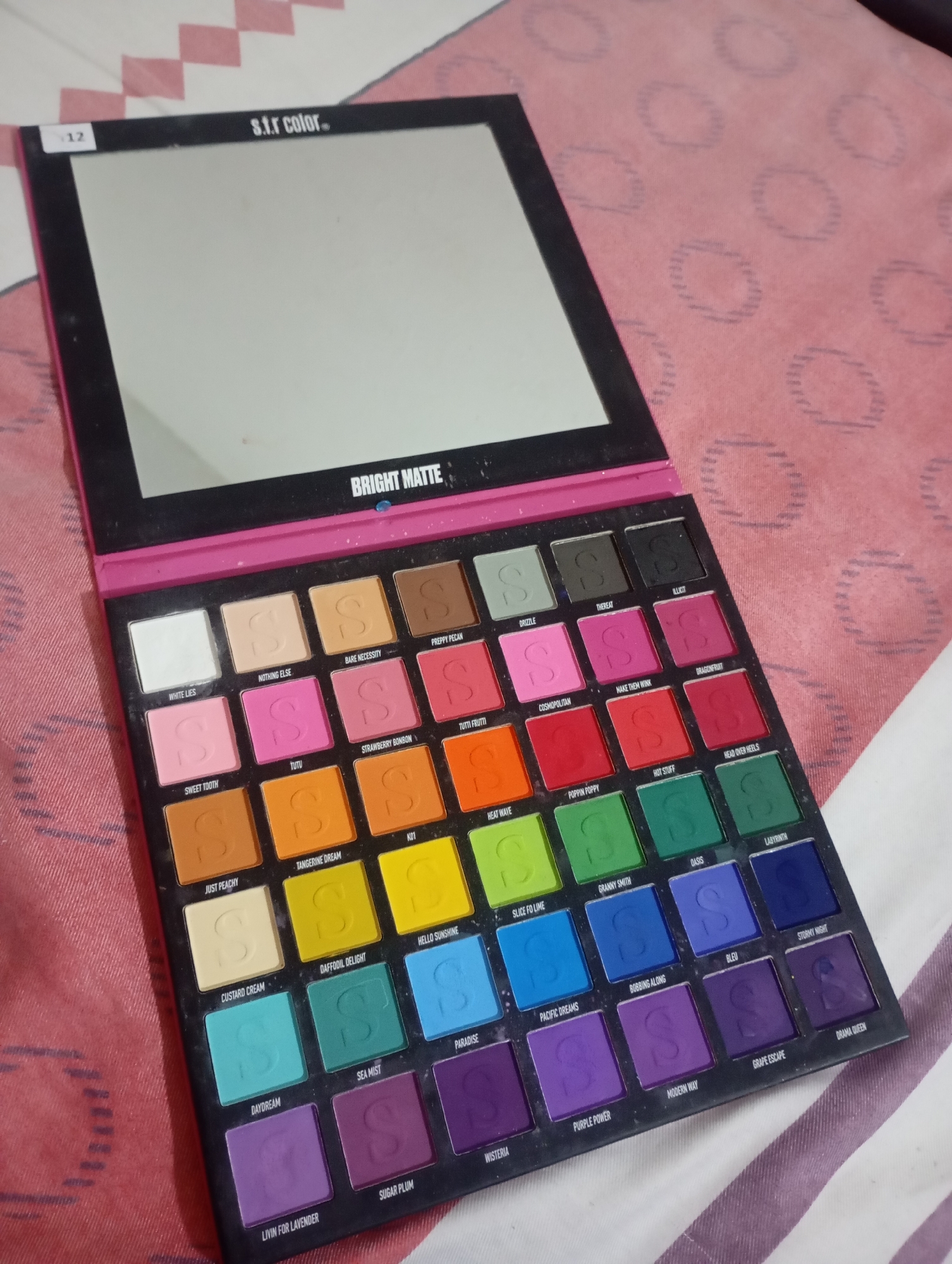 An open makeup palette with a large mirror is displayed on a patterned pink and white surface. The palette has a black casing with a pink interior. It contains 36 eyeshadow pans, each with an embossed "S" logo. The eyeshadow colors range from white and pastels to bright neons and deep jewel tones. The names of the eyeshadow shades are printed in small white text below each pan. The palette is positioned with its hinge on the left side. The background surface has a geometric diamond pattern.