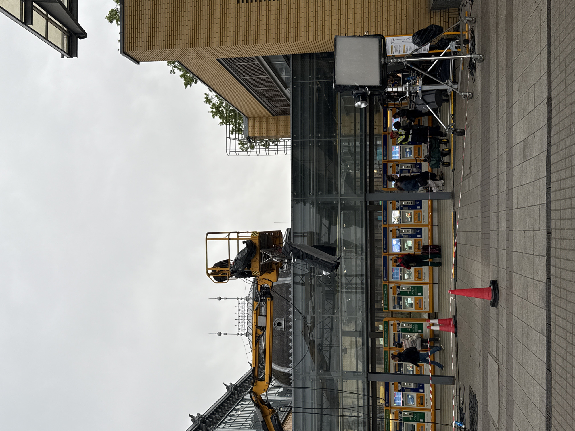 The image shows a scene in Budapest, Hungary, likely near a train station or public transport hub. A yellow hydraulic lift is positioned next to a modern glass-walled building, possibly for maintenance or construction. In the background, part of an older building with spires is visible. The area is pedestrianized, with tiled paving. A group of people are using what appear to be ticket machines or vending machines. Several orange traffic cones are placed on the pavement, possibly to cordon off an area. Behind the vending machine there are lighting equipments for an event/filming. The sky is overcast.
