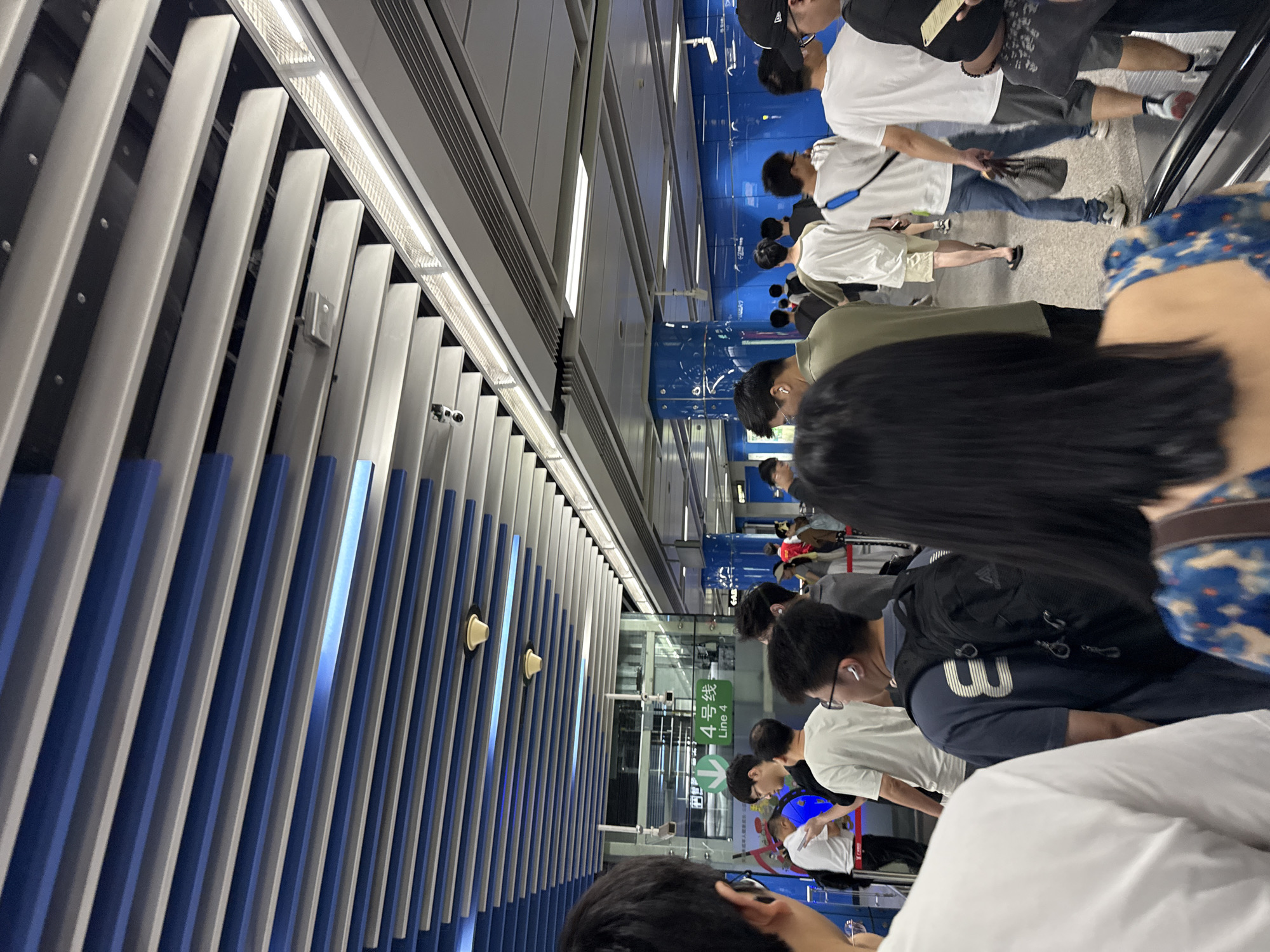 This image captures a scene at a subway station in Haizhu District, China. A diverse group of people are visible, mostly standing and walking, with many facing away from the camera. In the background, signage in Chinese indicates "4号线 Line 4" with a downward-pointing green arrow, suggesting this is a platform for that subway line. The environment features a modern, clean interior with a distinctive ceiling design composed of alternating blue and white slats. Fluorescent lighting is integrated into the ceiling, illuminating the area. Blue tiled pillars are also present, adding to the station's aesthetic. The overall atmosphere appears to be that of a busy transit hub, with individuals seemingly queuing or moving towards their desired destinations.
