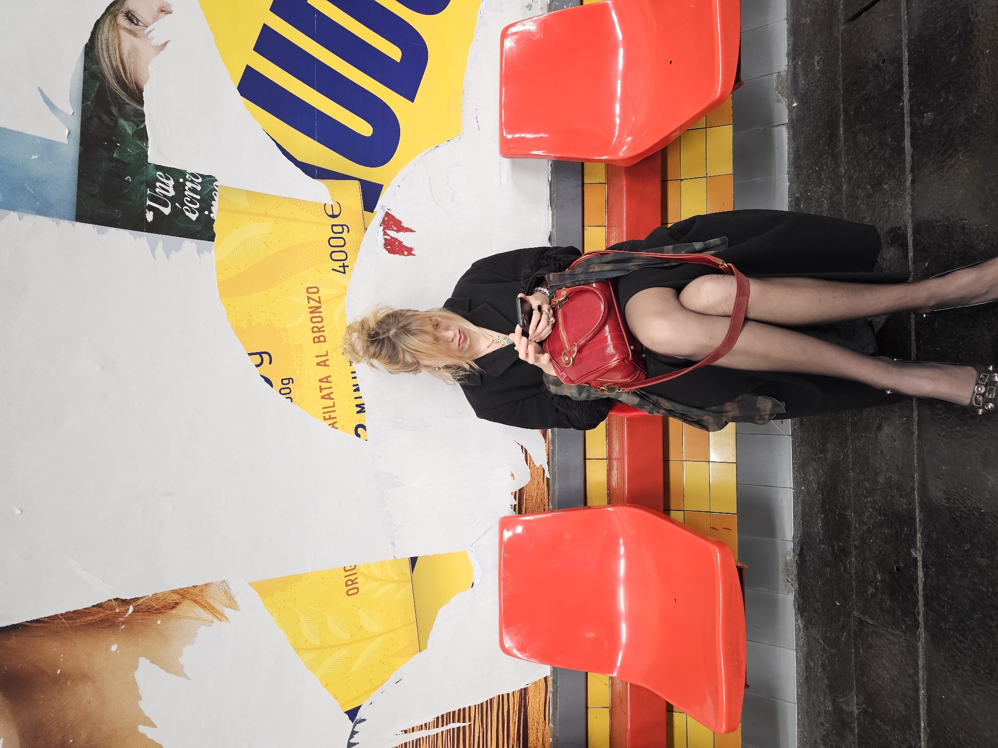 A woman sits on one of two red plastic seats in a Paris Metro station, using a smartphone. The flooring is tiled in yellow and orange; beside this are gray tiles. The floor beyond the seat is a dark gray. Behind the seating is a wall plastered with layers of torn posters. The visible posters contain text in French and images of people. The woman has blonde hair pulled into a loose bun. She wears a black overcoat and what appear to be black tights and silver pointed-toe heels, and a red leather purse rests on her lap. Her legs are crossed.