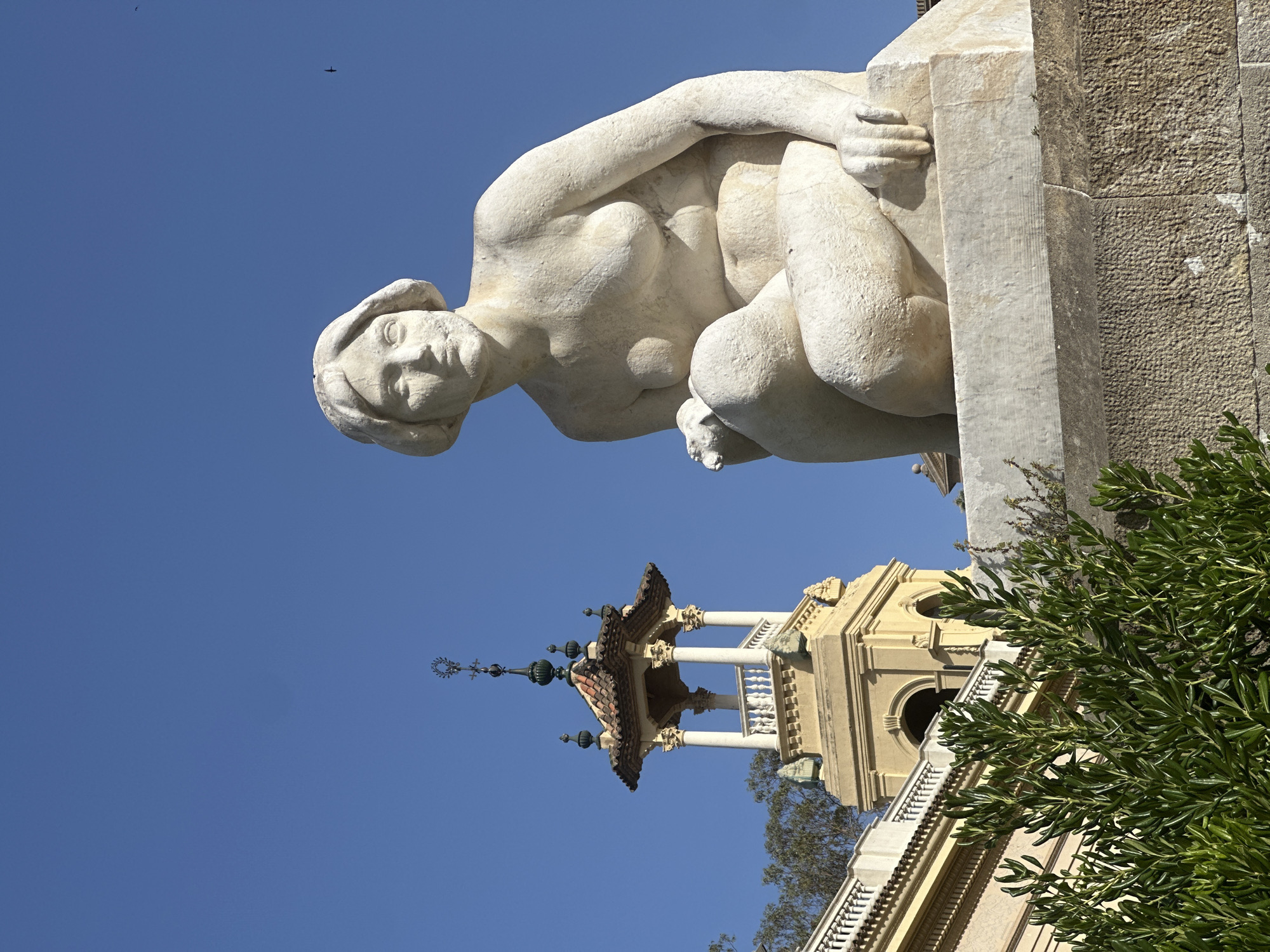 The image shows a white marble statue of a seated woman with her legs crossed and her left hand resting on her lap. The statue is positioned in front of a building with a tower, and lush green foliage is visible in the foreground. The sky is clear and blue. The statue and the architecture suggest the location is the Park Güell in Barcelona, Spain, which is known for its distinctive architectural elements and artistic sculptures. The statue appears to be a classical-style representation of a female figure, likely a decorative element within the park. The building in the background is an ornate structure with a tiled roof and arched openings, characteristic of Antoni Gaudí's architectural style, which dominates Park Güell. The green branches in the foreground are part of the park's landscaping, contributing to the natural and artistic ambiance.