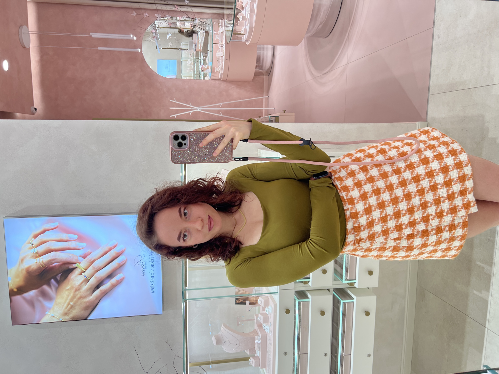 A young woman with curly brown hair takes a selfie in a brightly lit jewelry store. She is wearing a long-sleeved olive green top and a textured orange and white checkered skirt. Her phone, in a sparkly pink case with a pink strap, is held up to take the photo. Behind her, jewelry is displayed in glass cases, and the walls are painted in shades of pink and grey. The overall ambiance suggests a modern and chic retail environment.